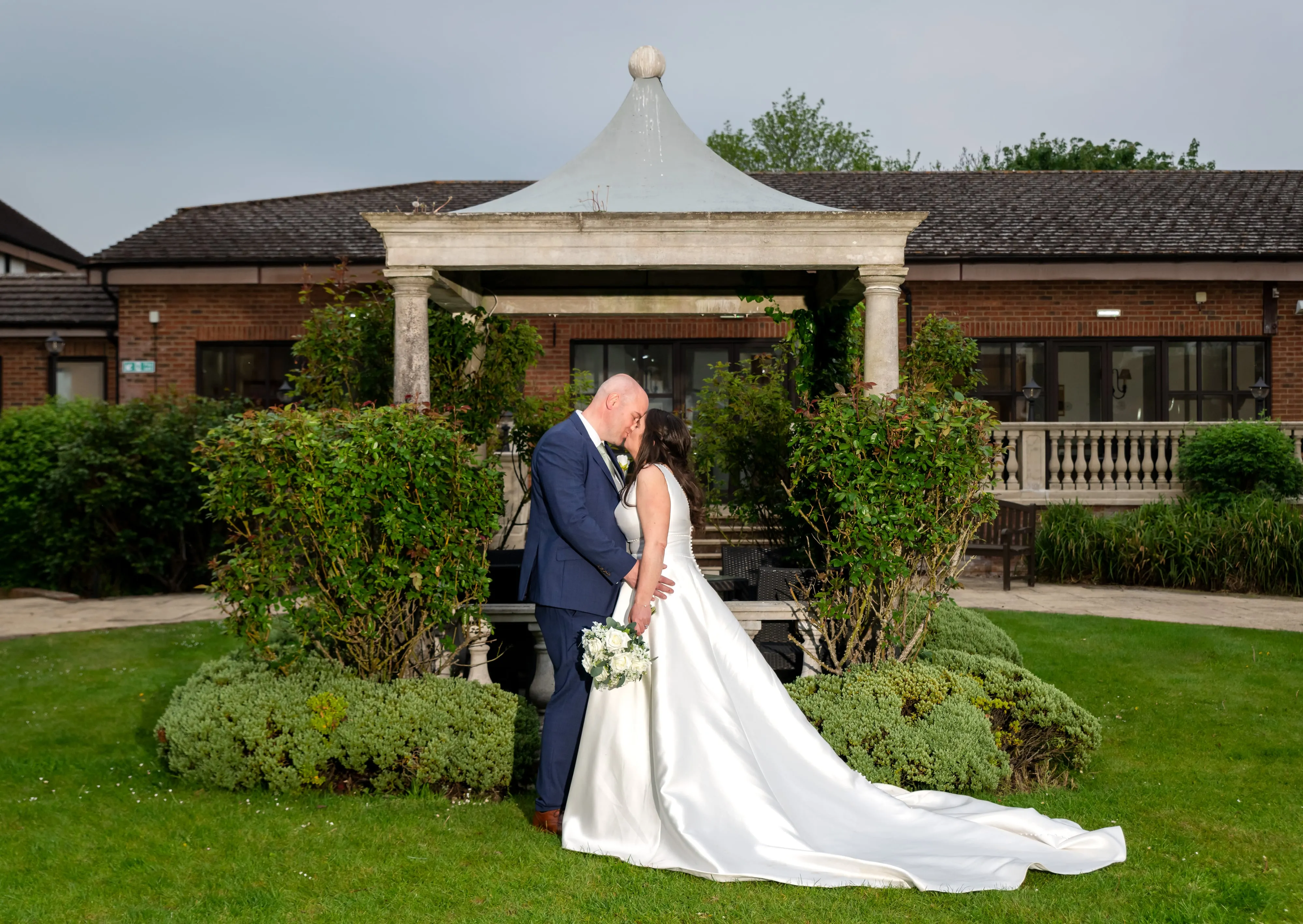 Bride and groom kiss beneath the stone pavilion in the Belfry gardens, full-length silhouette with manicured shrubs flanking