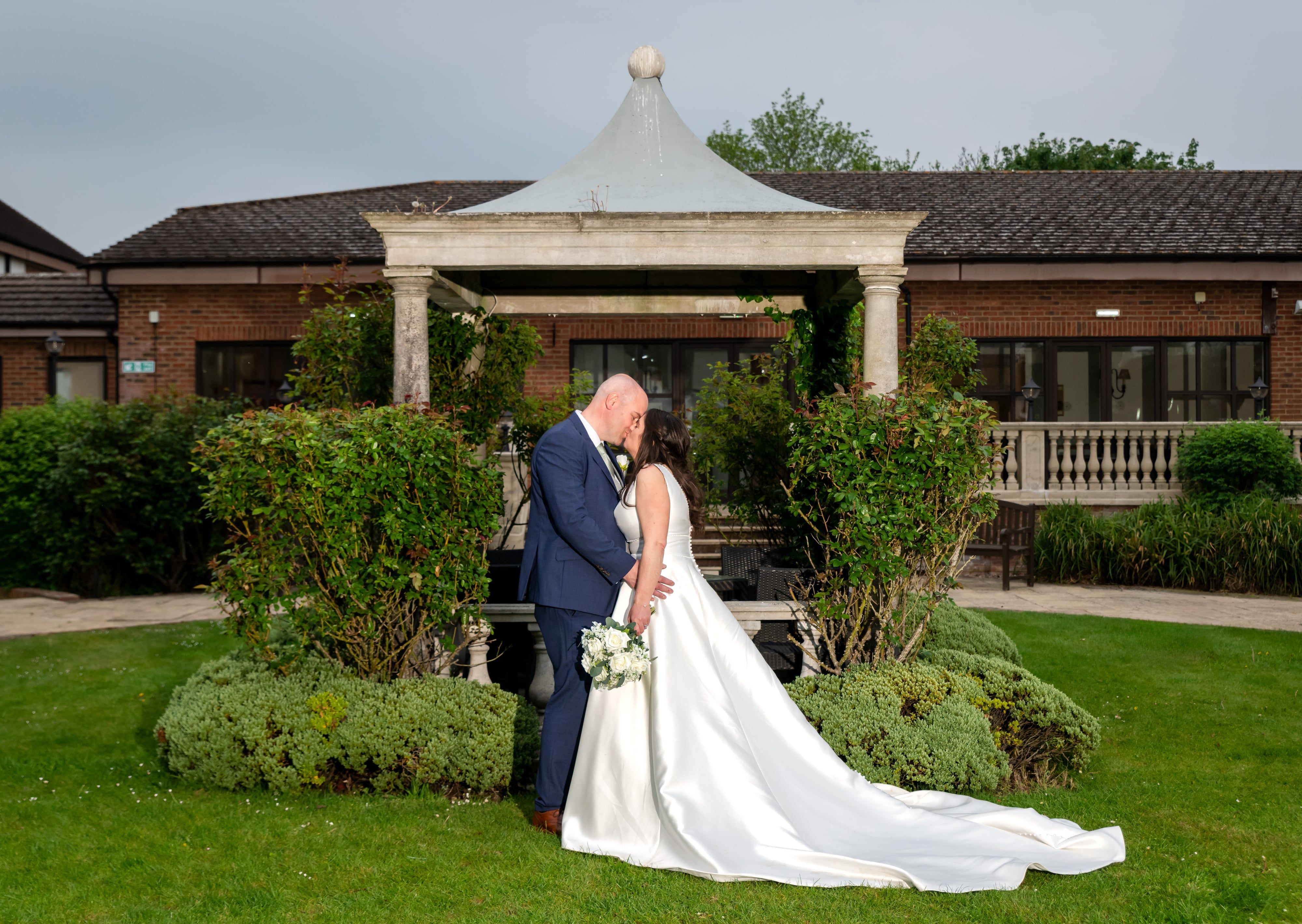 Bride and groom kiss beneath the stone pavilion in the Belfry gardens, full-length silhouette with manicured shrubs flanking