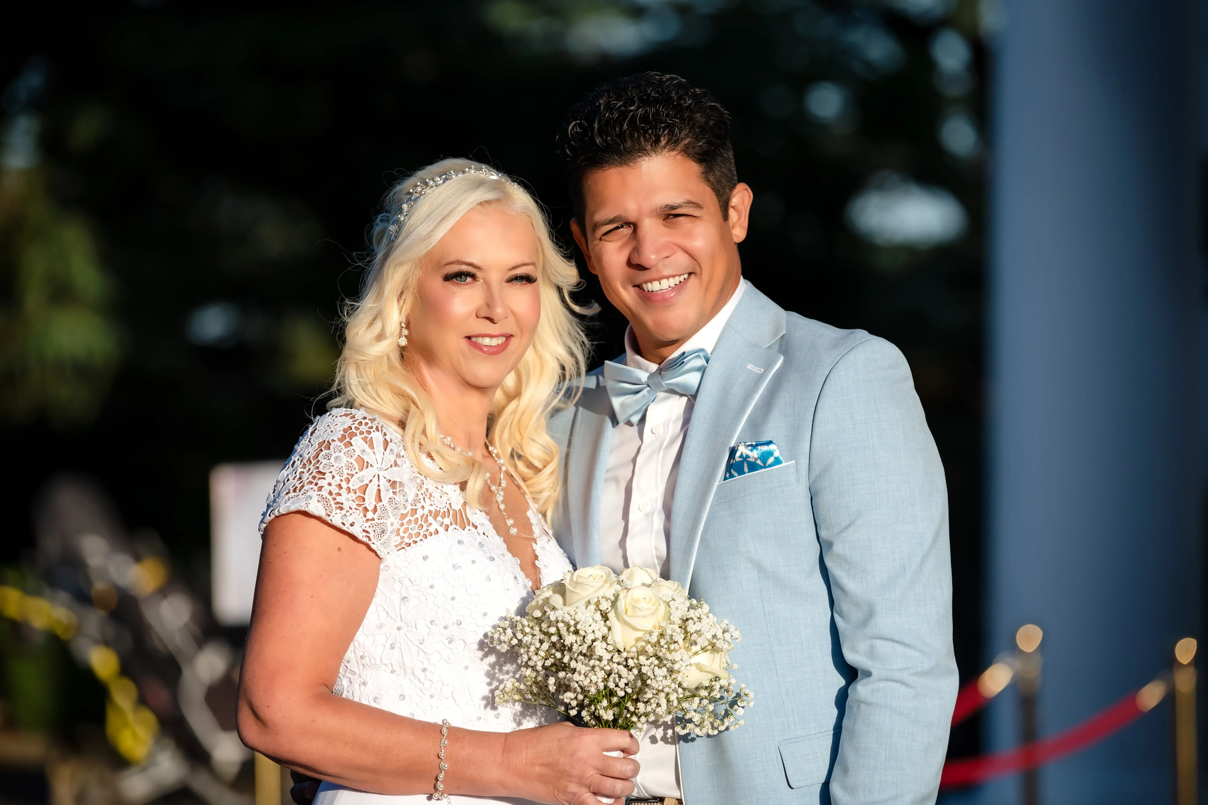 Smiling close-up portrait of bride and groom outside Dyrham Park, bride with diamanté headpiece, groom in pale-blue suit and bow tie