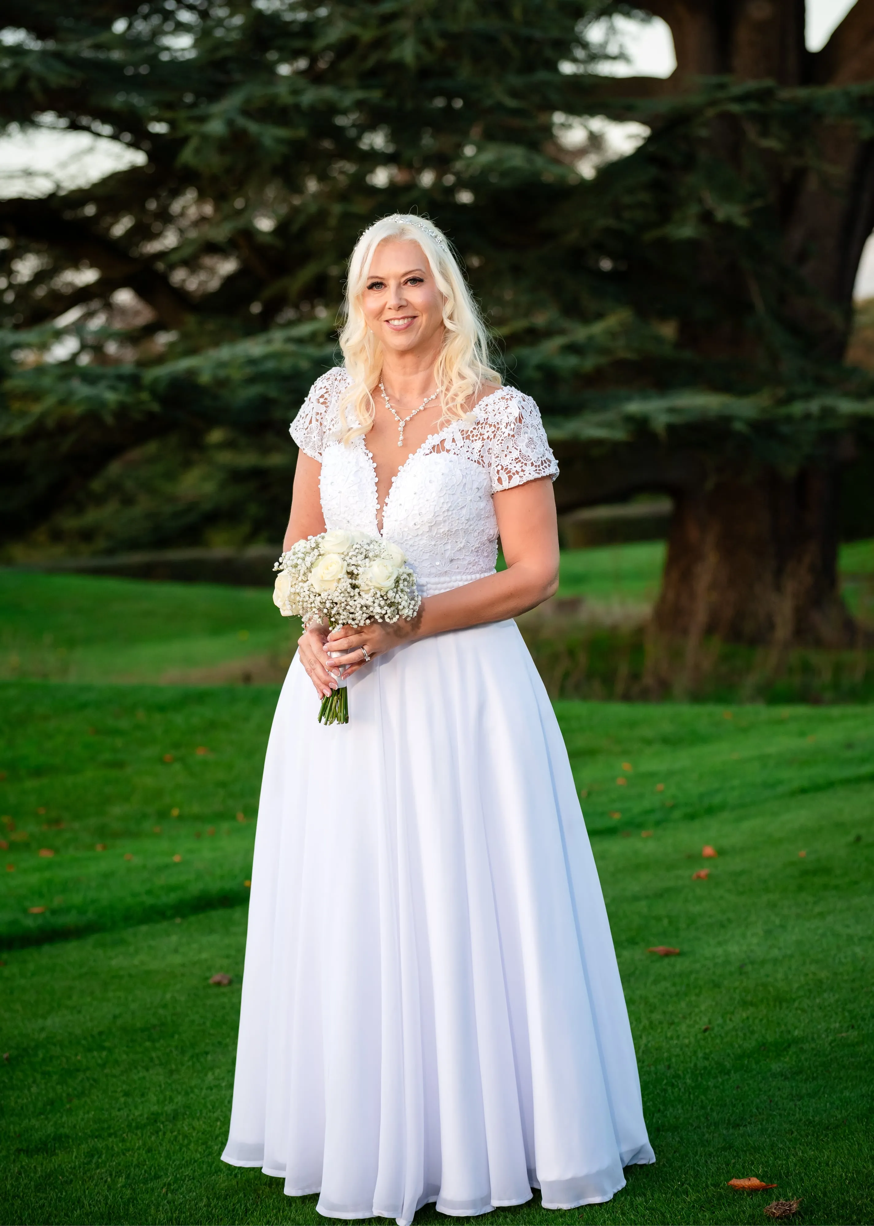 Solo bride portrait under the Dyrham Park cedar tree, full-length white lace gown holding cream-rose bouquet