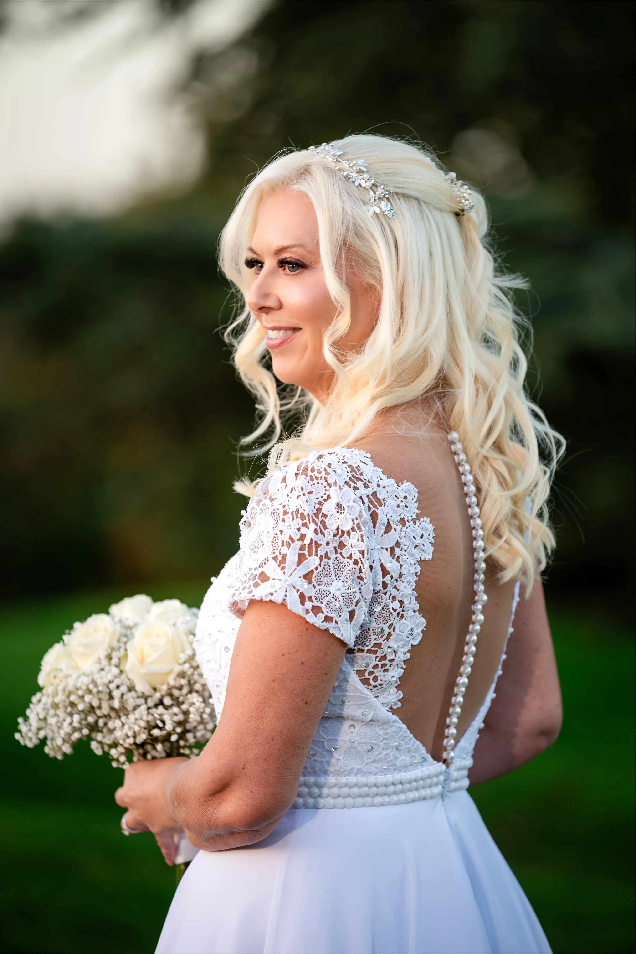 Bride seen from behind with half-up blonde hair, white lace cap-sleeve gown with vertical pearl-button back detail
