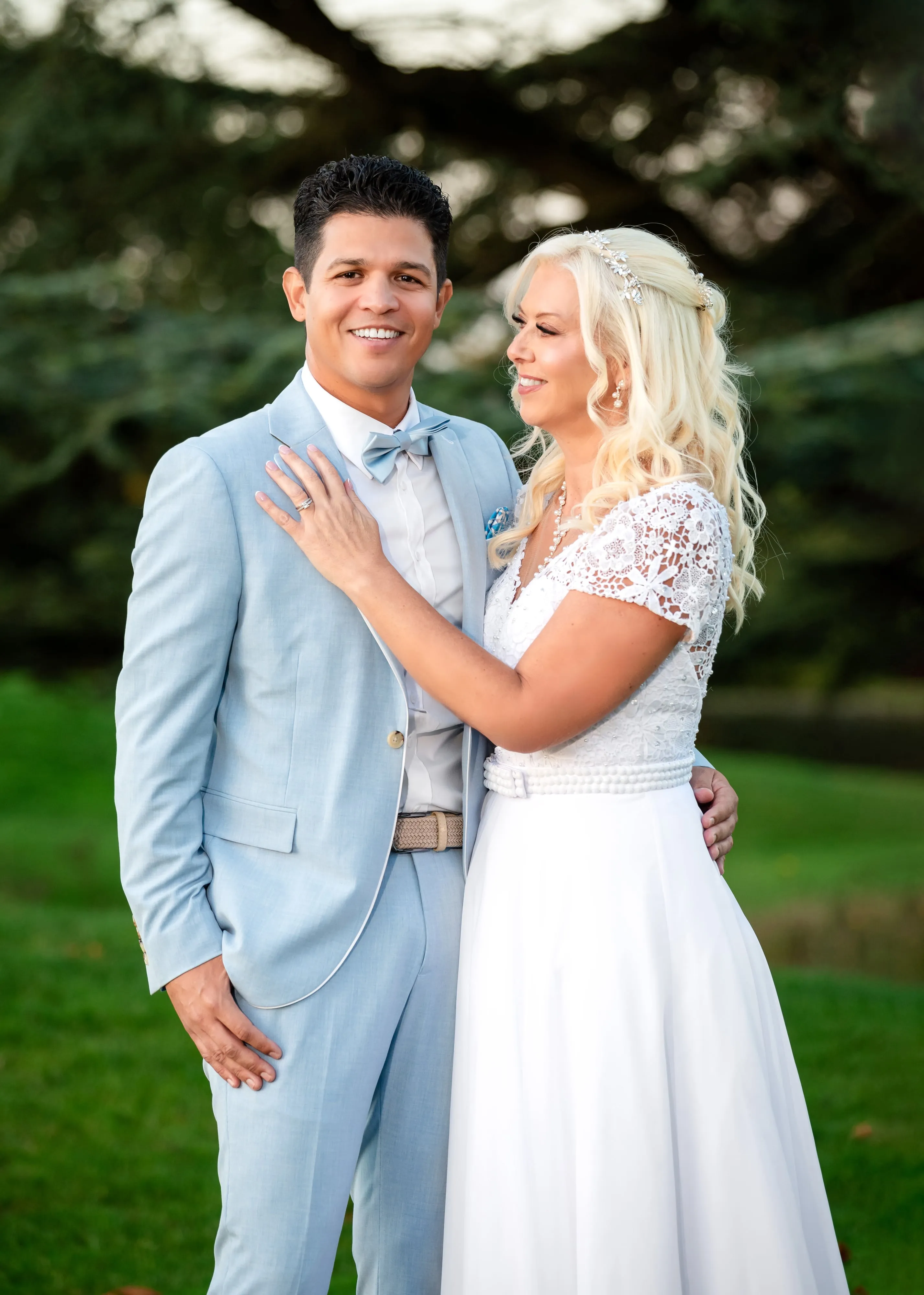 Close-up couple portrait, bride's hand on groom's chest with wedding ring visible, cedar-lined parkland behind