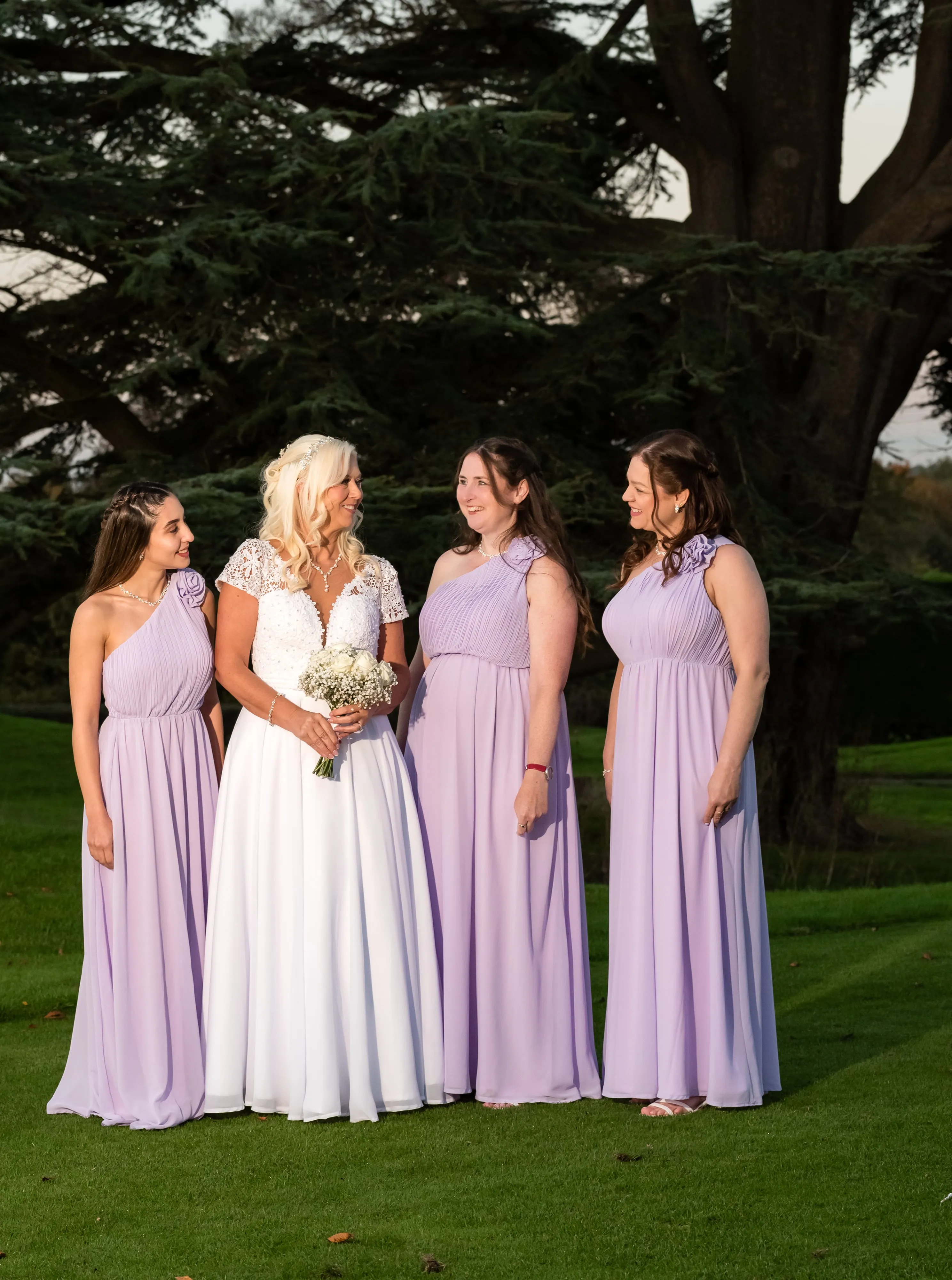 Bride laughs with three bridesmaids in lilac one-shoulder dresses under the Dyrham Park cedar tree