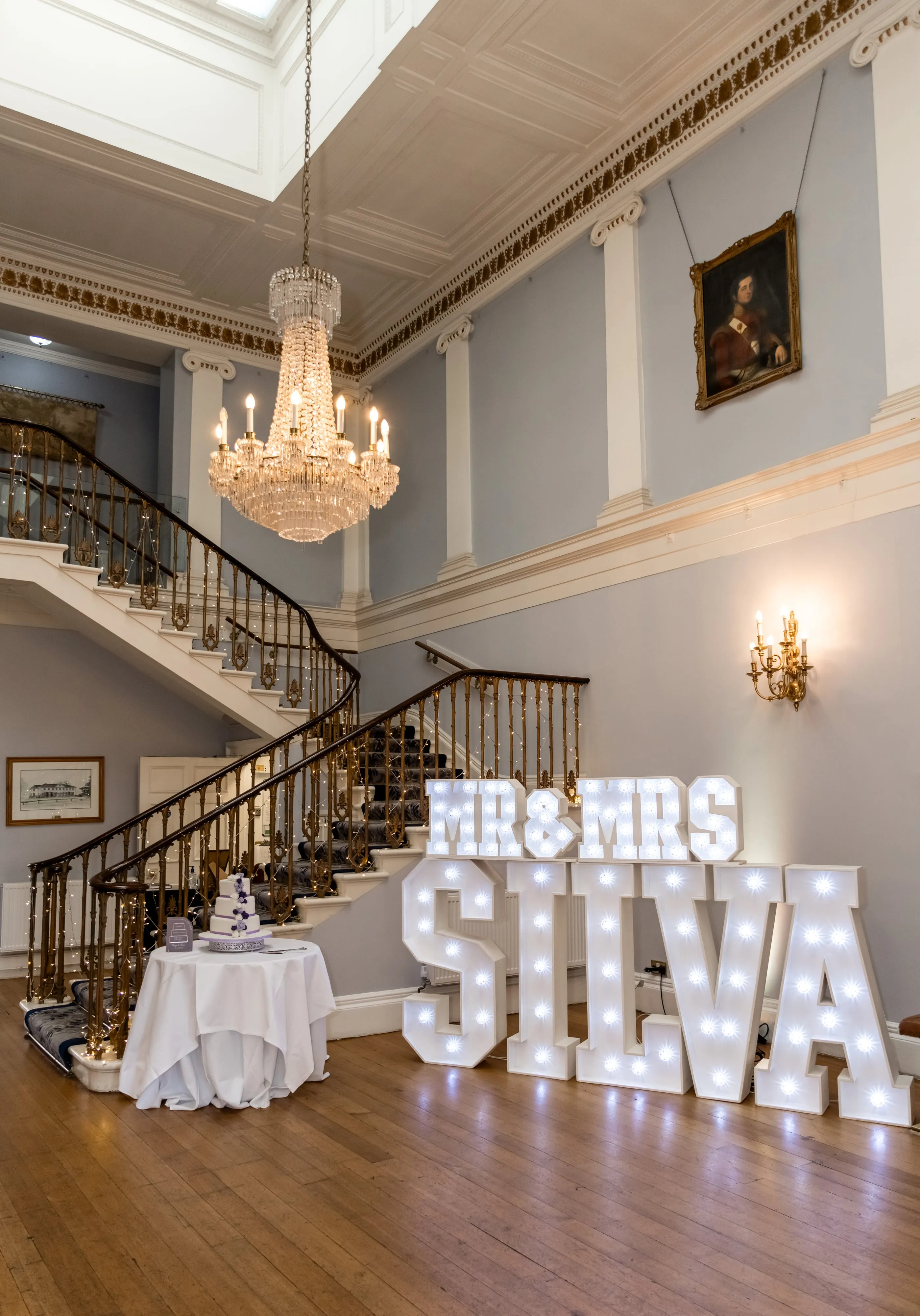 Grand Dyrham Park foyer dressed for the wedding — crystal chandelier, sweeping iron staircase, illuminated 'MR & MRS SILVA' marquee letters beside a three-tier cake