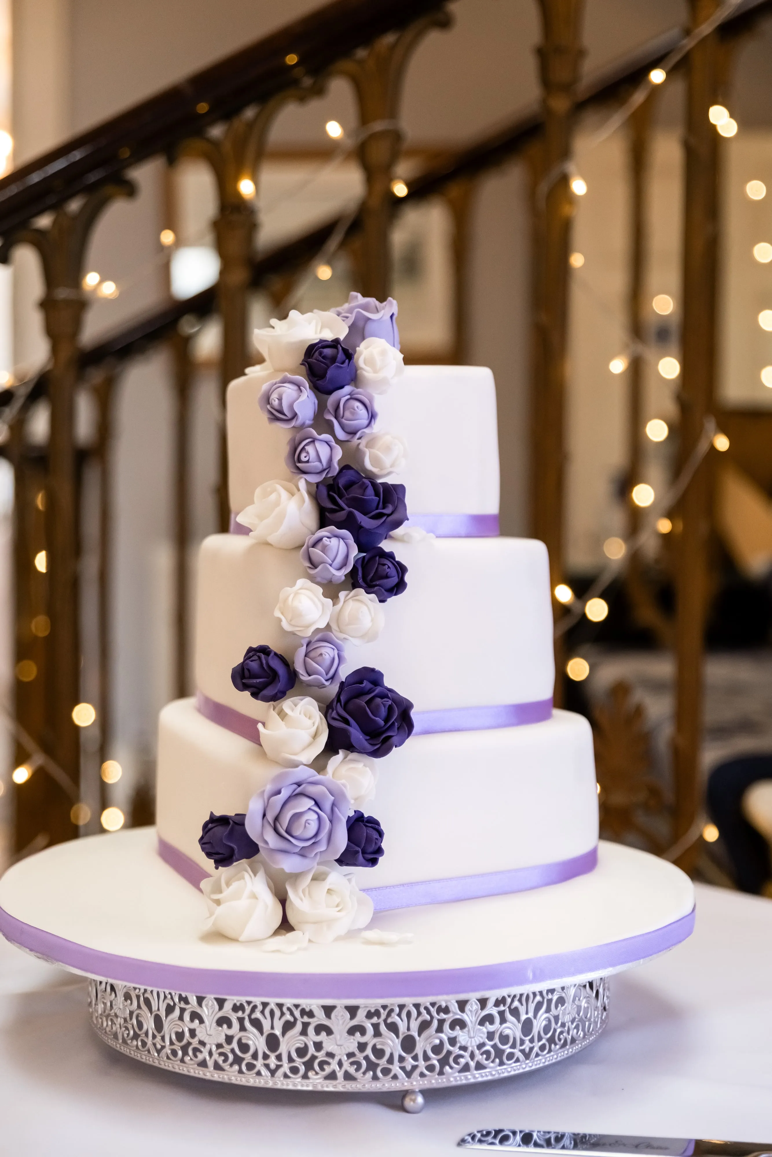 Three-tier white wedding cake with cascading purple and white roses, on a silver stand against a fairy-lit balustrade staircase