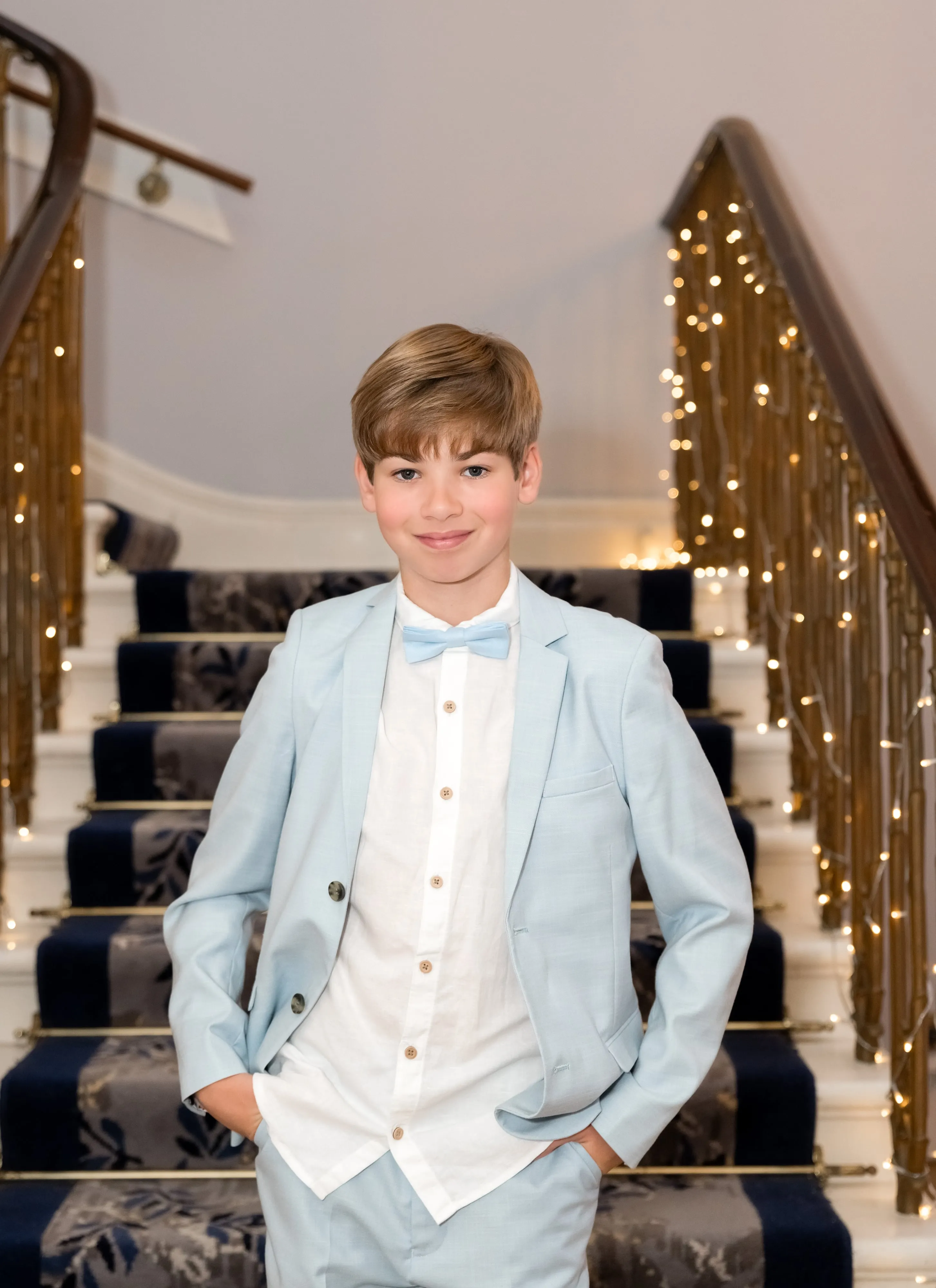 Young guest in pale-blue suit and matching bow tie stands on the Dyrham Park grand staircase, fairy-lit banister behind