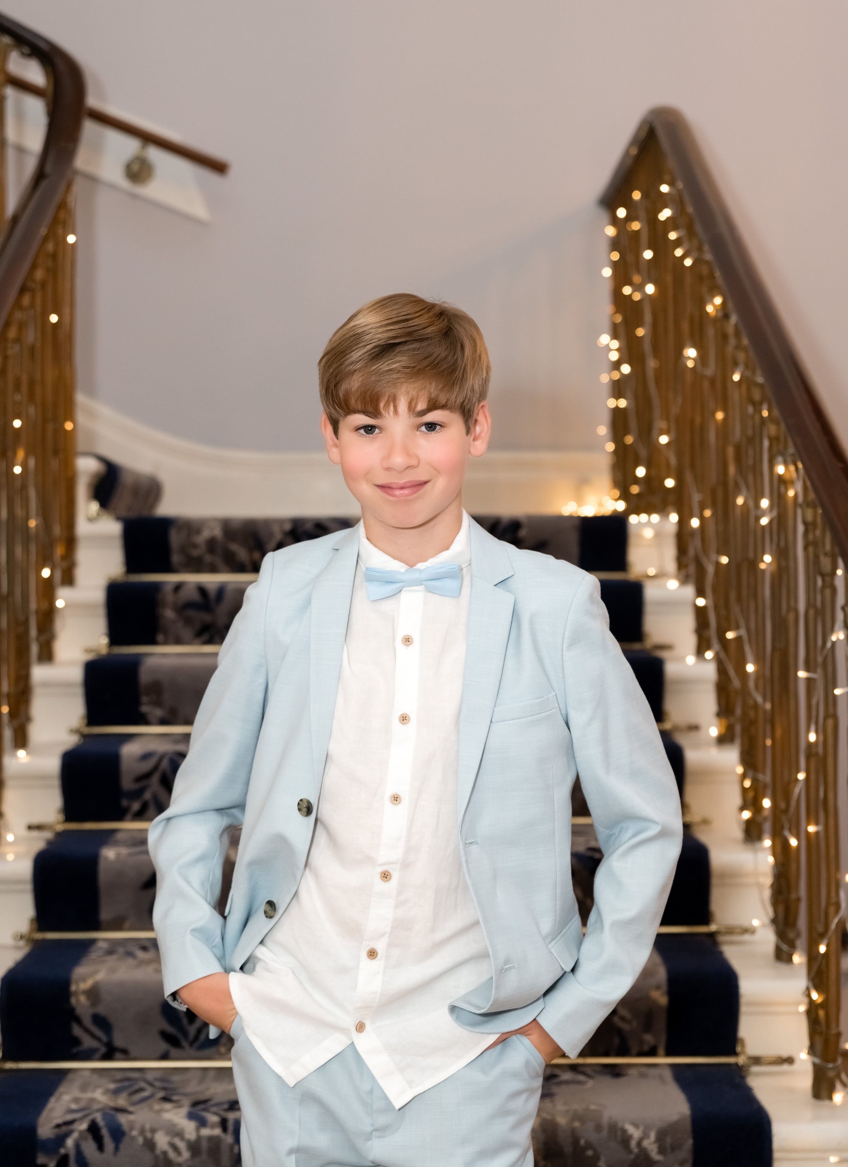 Young guest in pale-blue suit and matching bow tie stands on the Dyrham Park grand staircase, fairy-lit banister behind