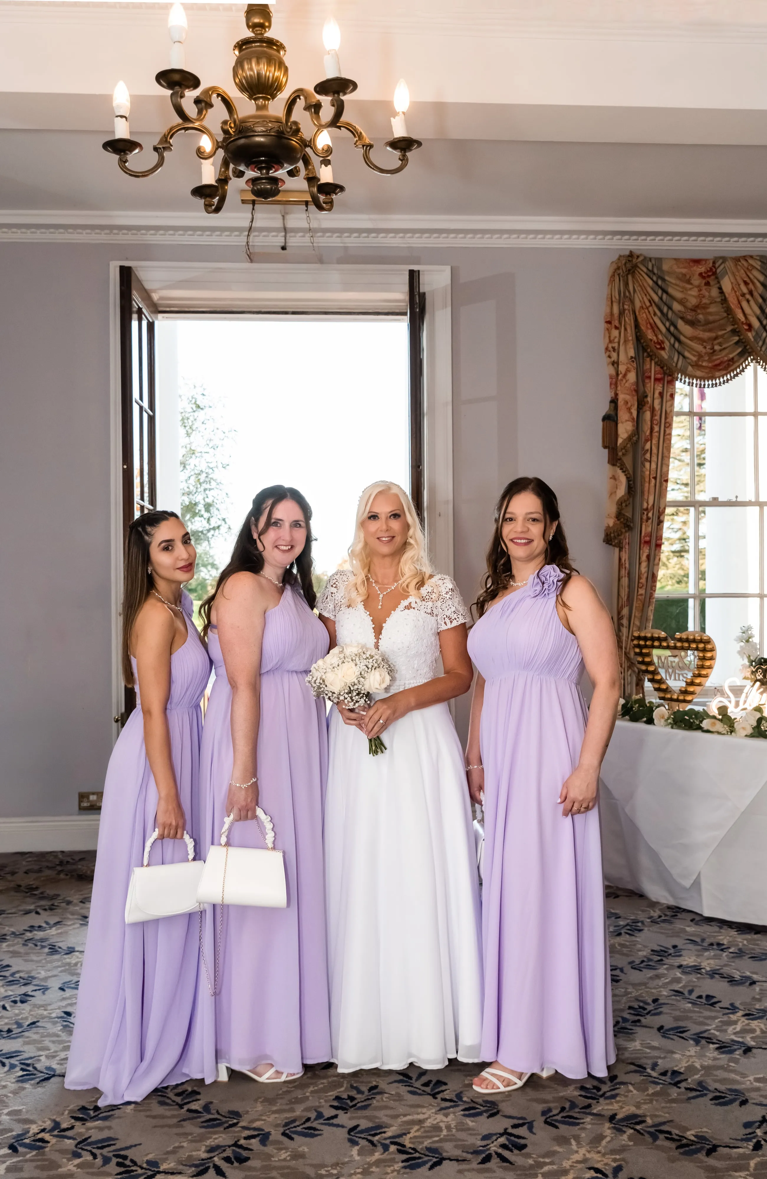 Bride and three bridesmaids in lilac dresses pose under a brass chandelier in the Dyrham Park drawing room
