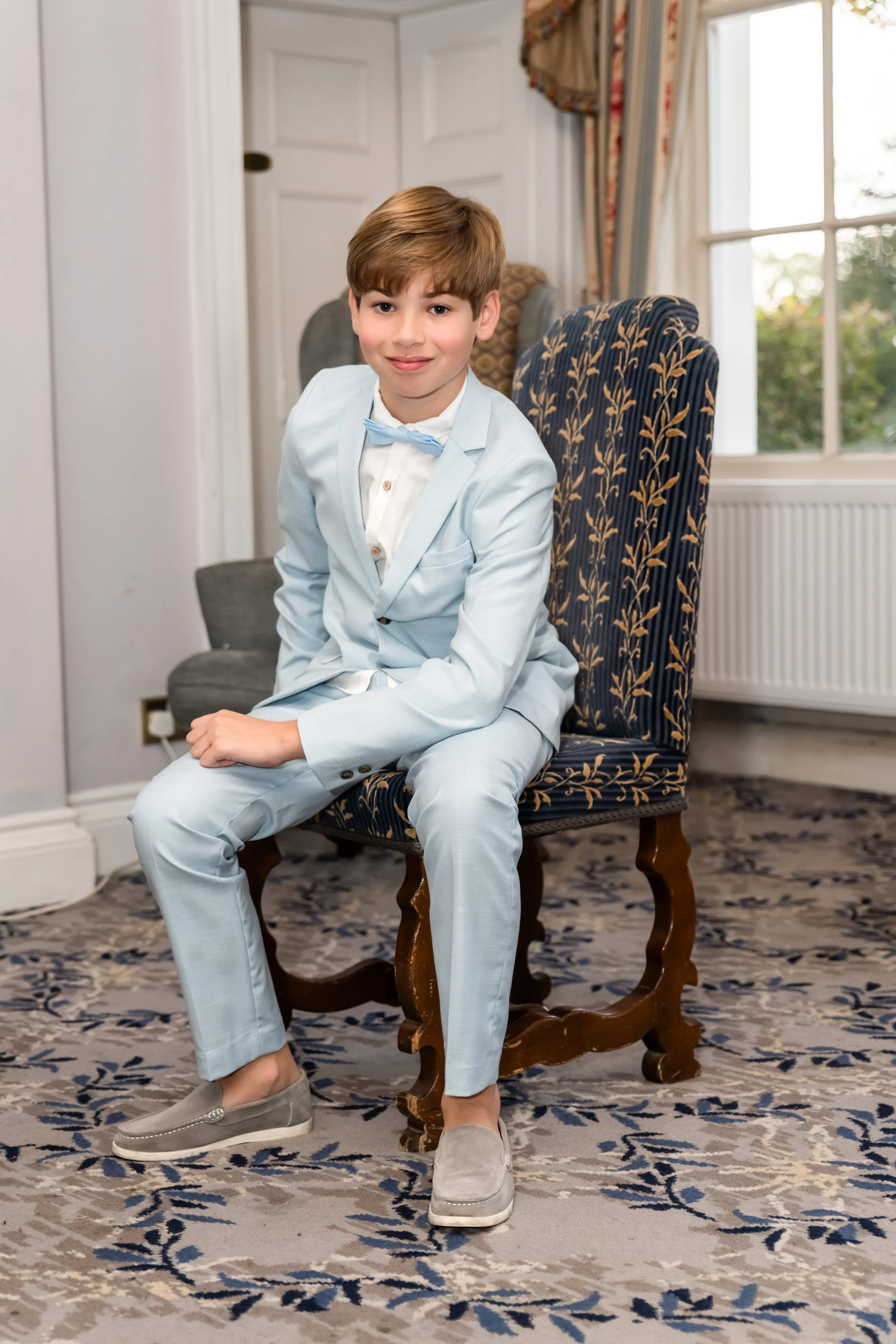 Young guest in pale-blue suit and bow tie sits on a navy-and-gold patterned wing chair in a Dyrham Park lounge