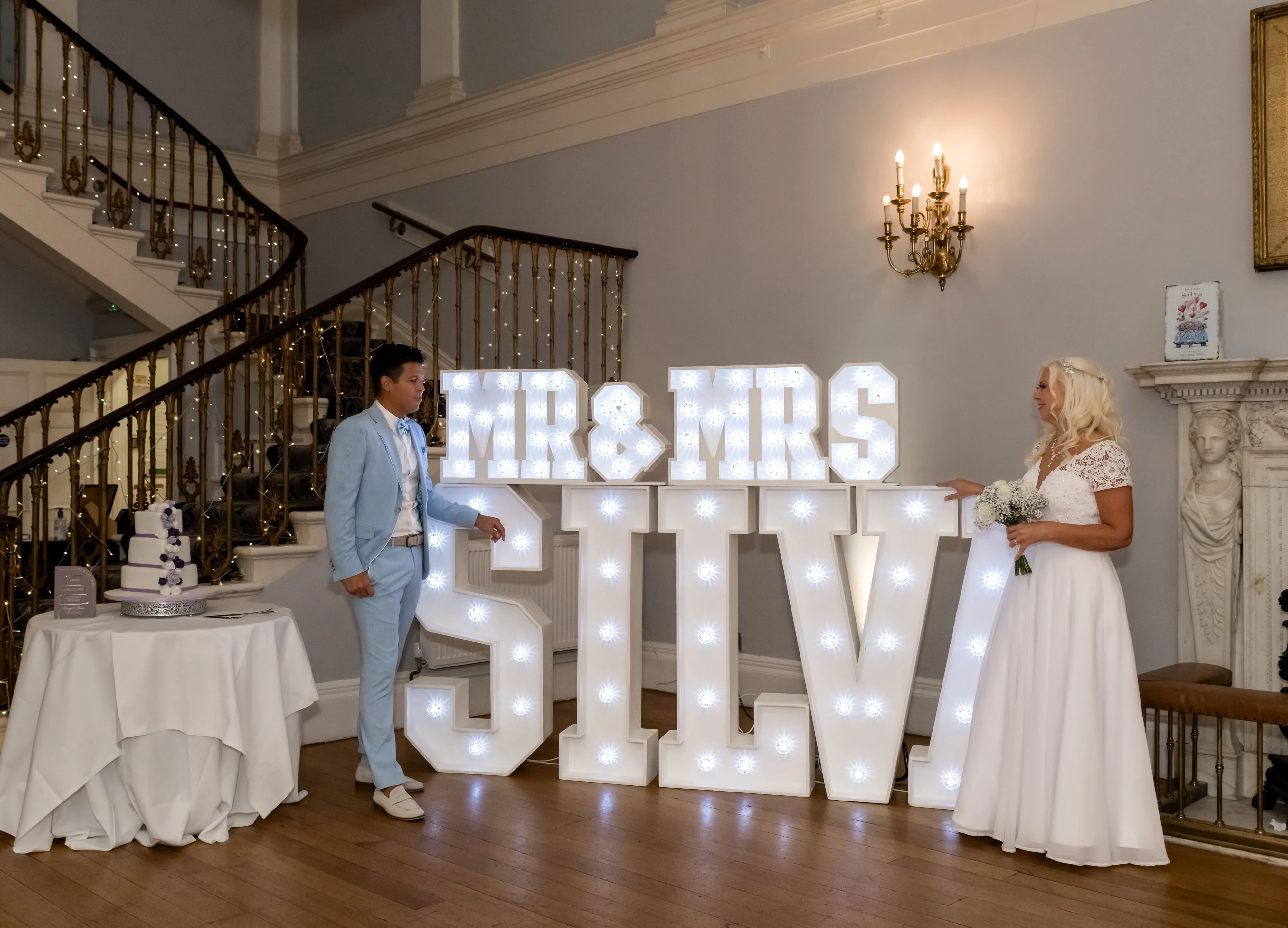 Bride and groom flank giant illuminated 'MR & MRS SILVA' marquee letters in the Dyrham Park foyer, three-tier cake to the left