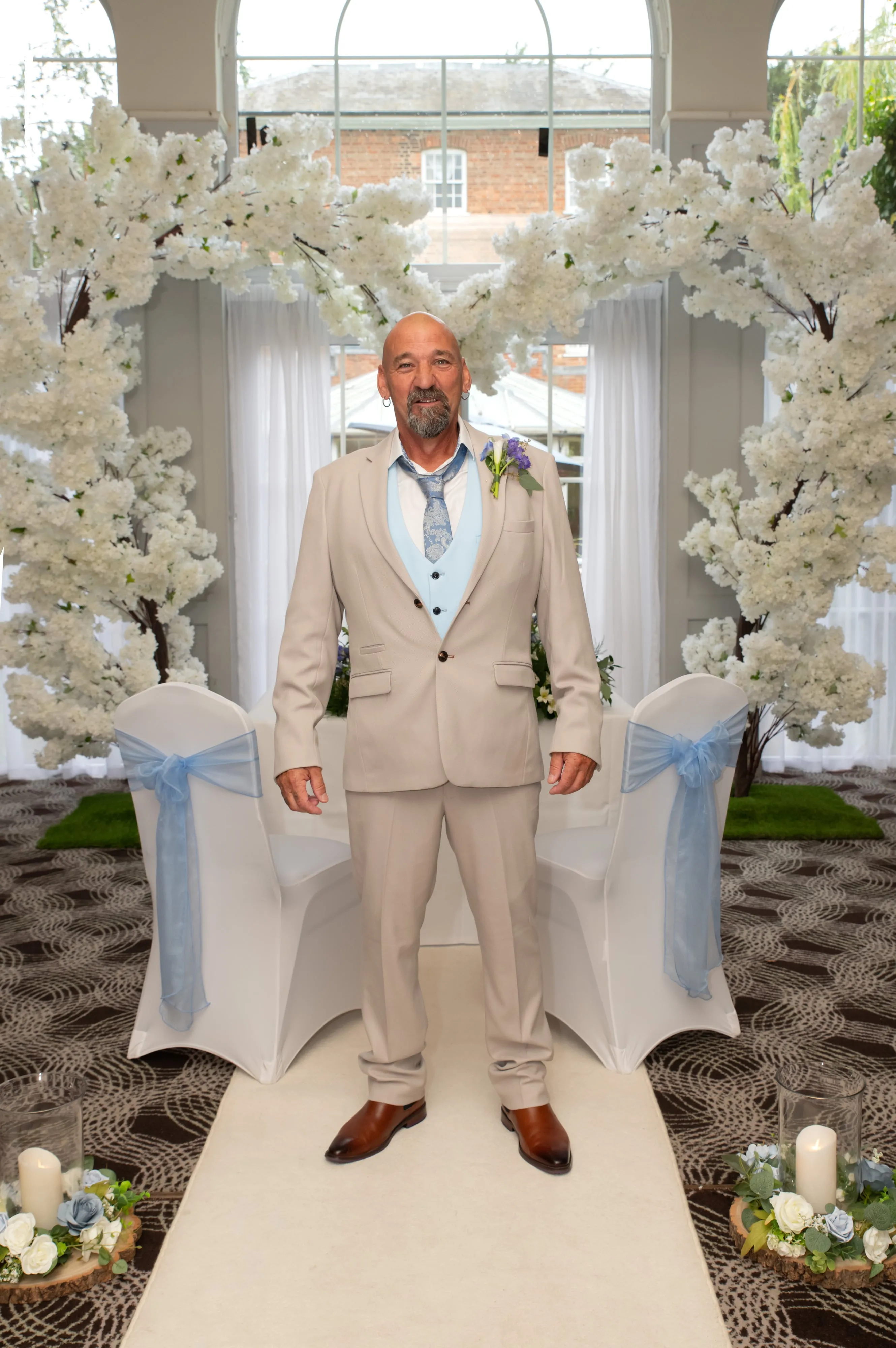 Father of the bride beneath the white floral ceremony arch in the Lovett Conservatory, blue-sash chairs flanking the aisle