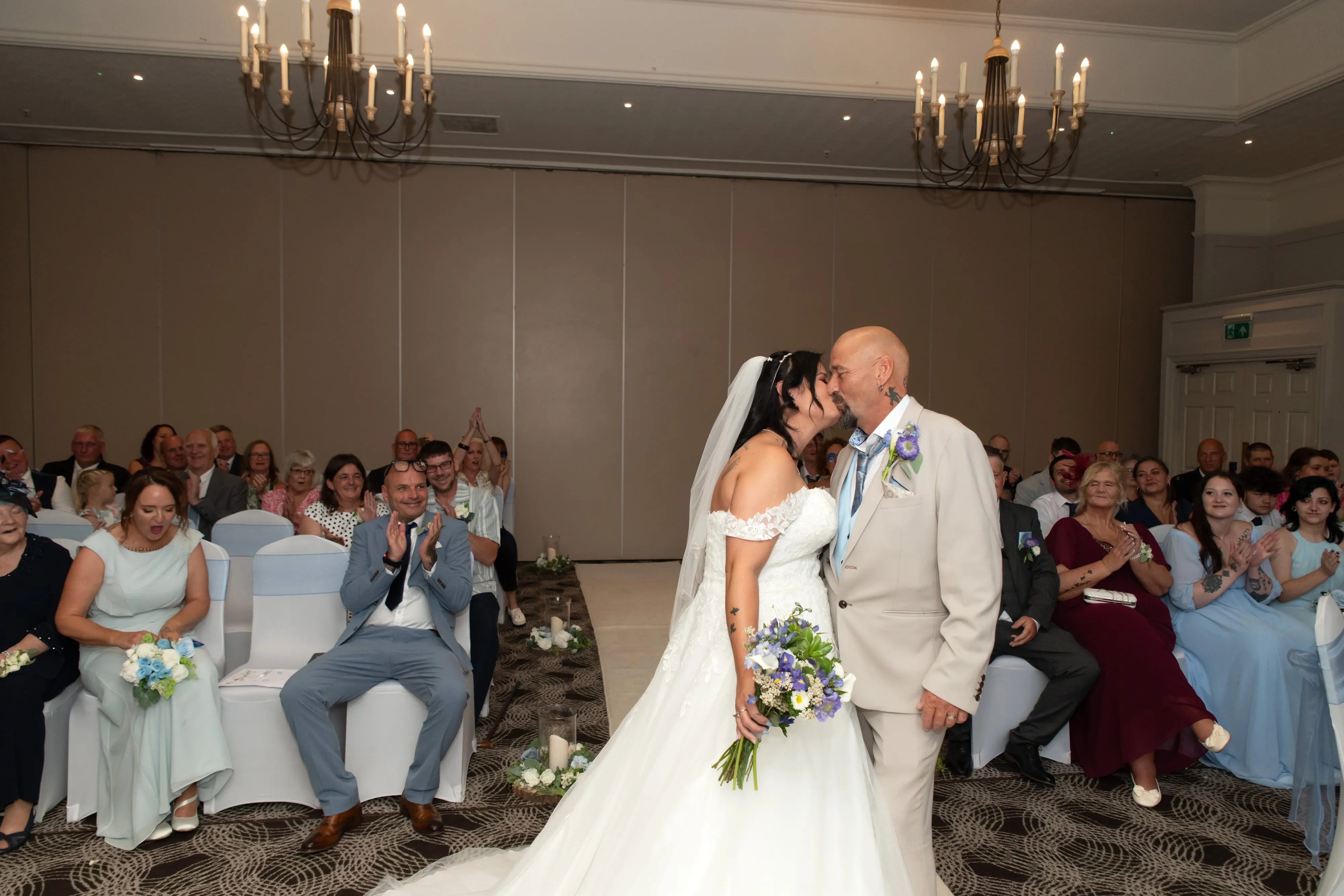 Bride and groom share their first kiss in the aisle as guests applaud beneath chandelier-lit ceremony room