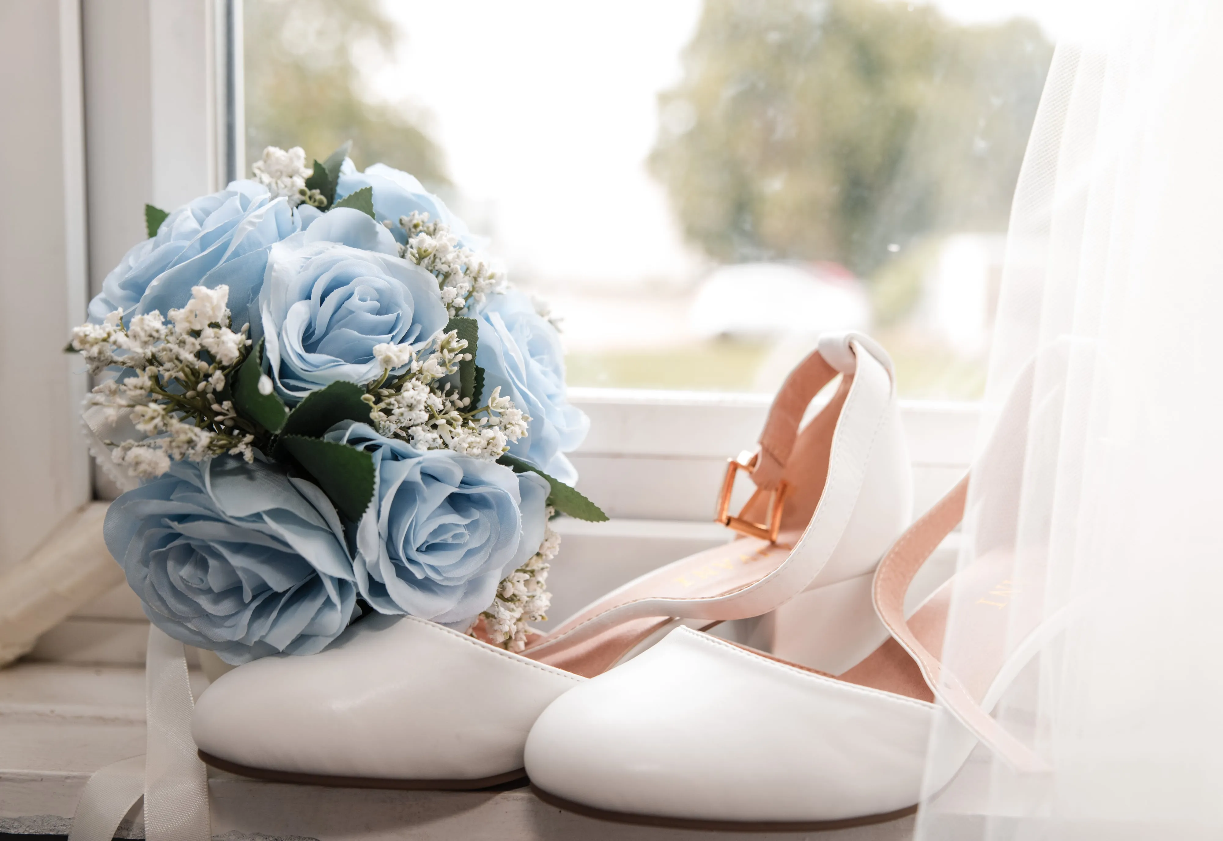 Bridal heels and a pale-blue rose bouquet rest on a sunlit windowsill before the ceremony