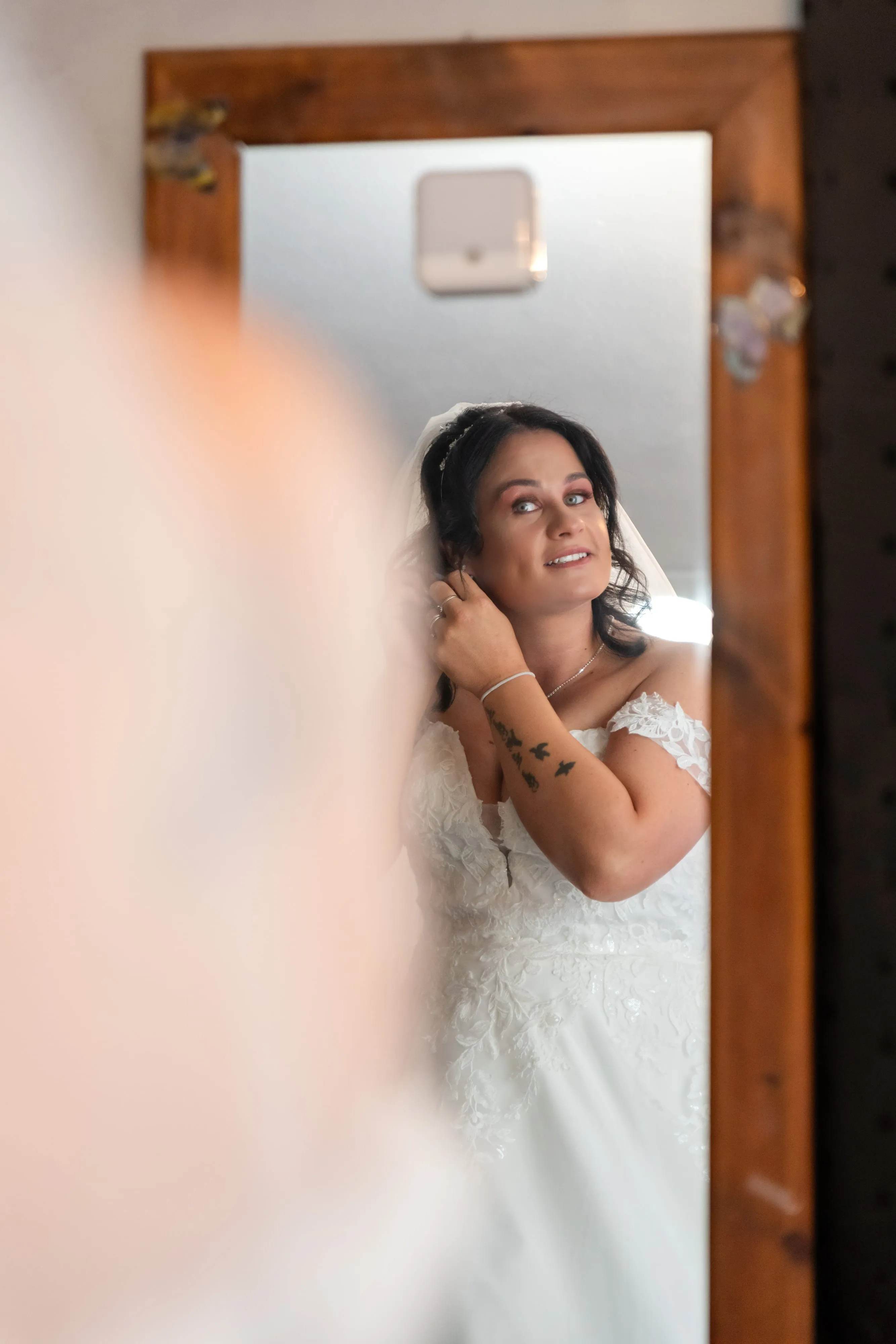 Bride adjusts her veil in a wood-framed mirror during getting-ready, soft shoulder-blur foreground
