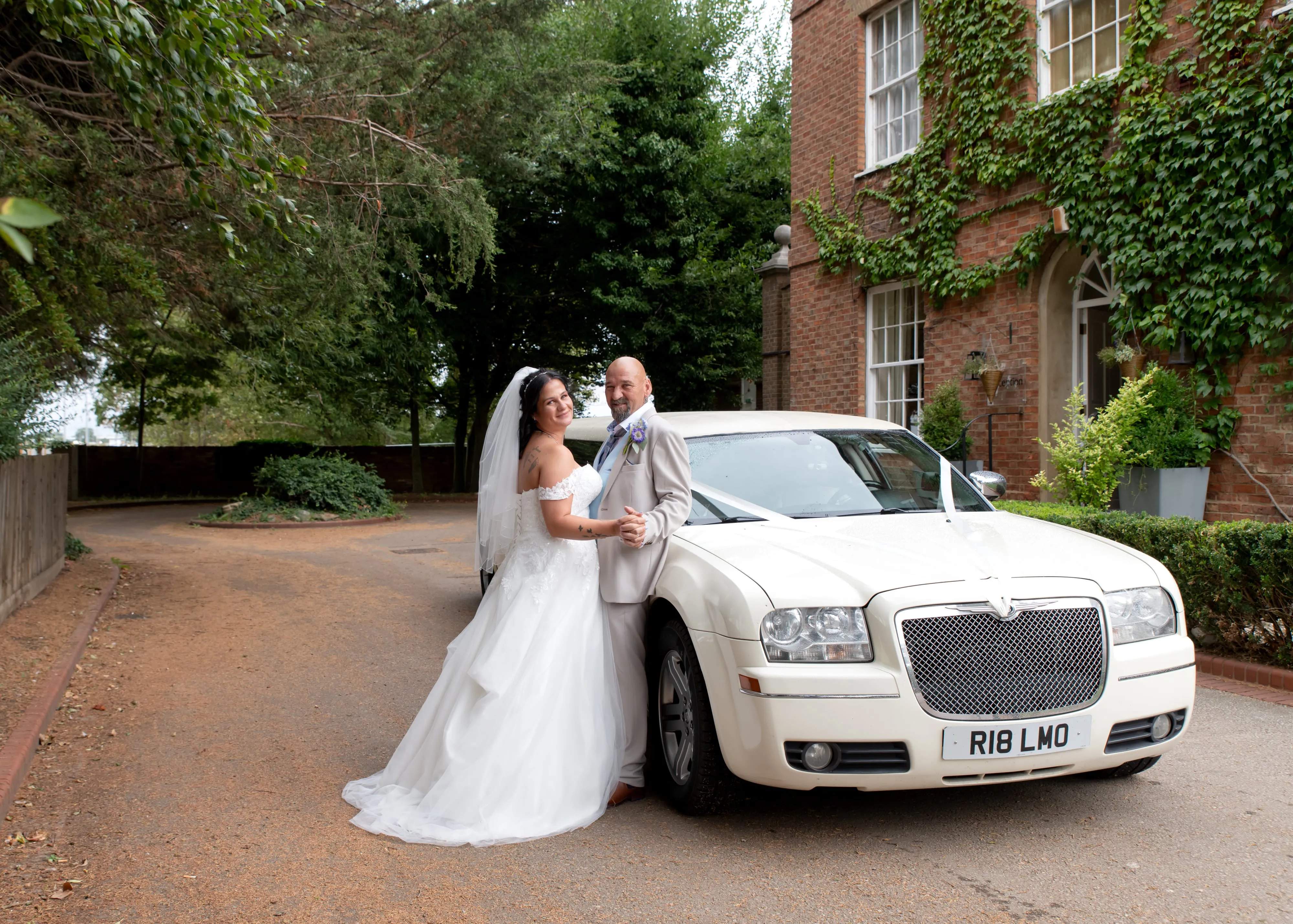 Bride and groom pose beside a white stretch limo on the ivy-covered red brick driveway