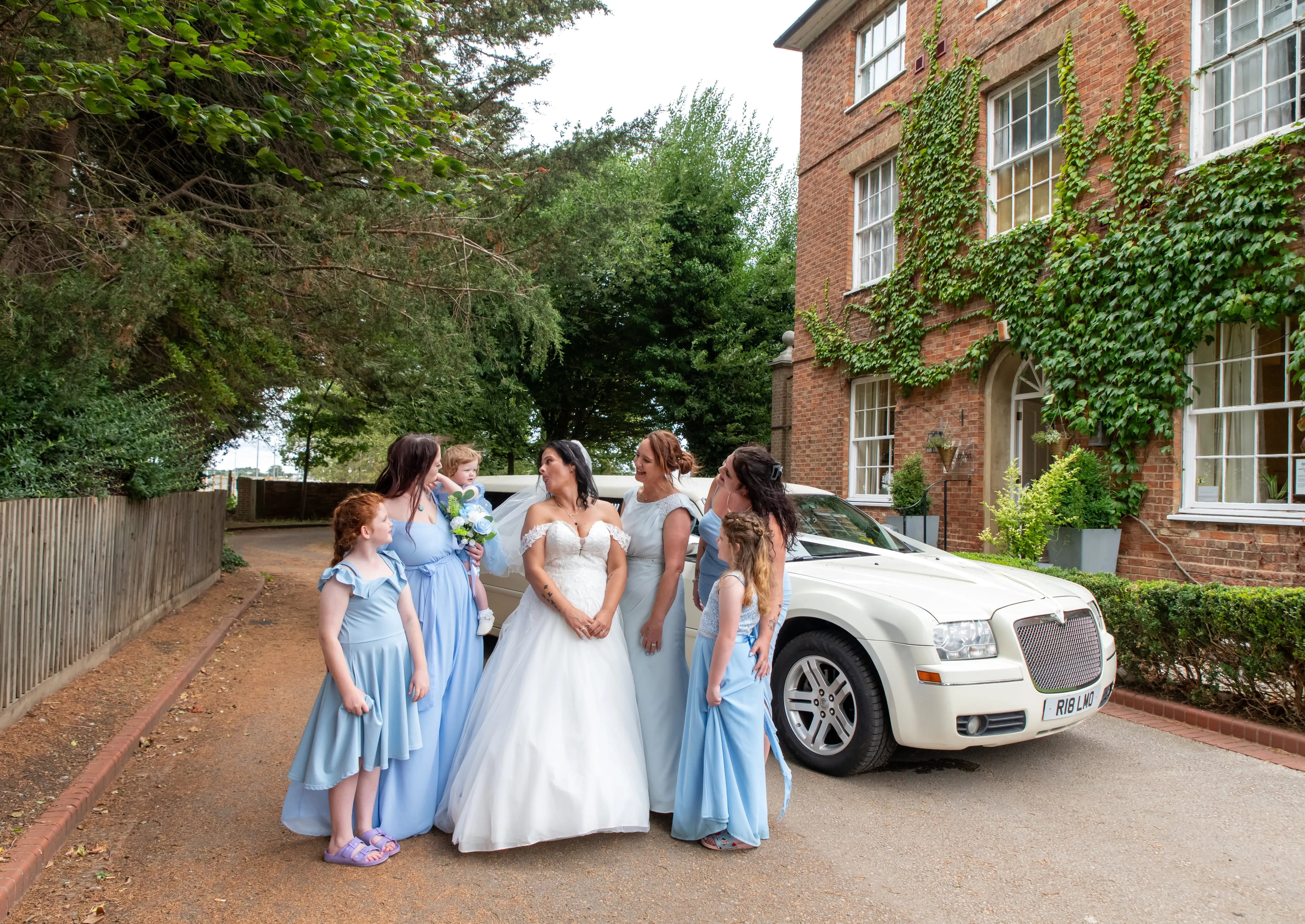 Bridal party in pale-blue dresses gather around the bride and groom by a white limo at the ivy-clad venue