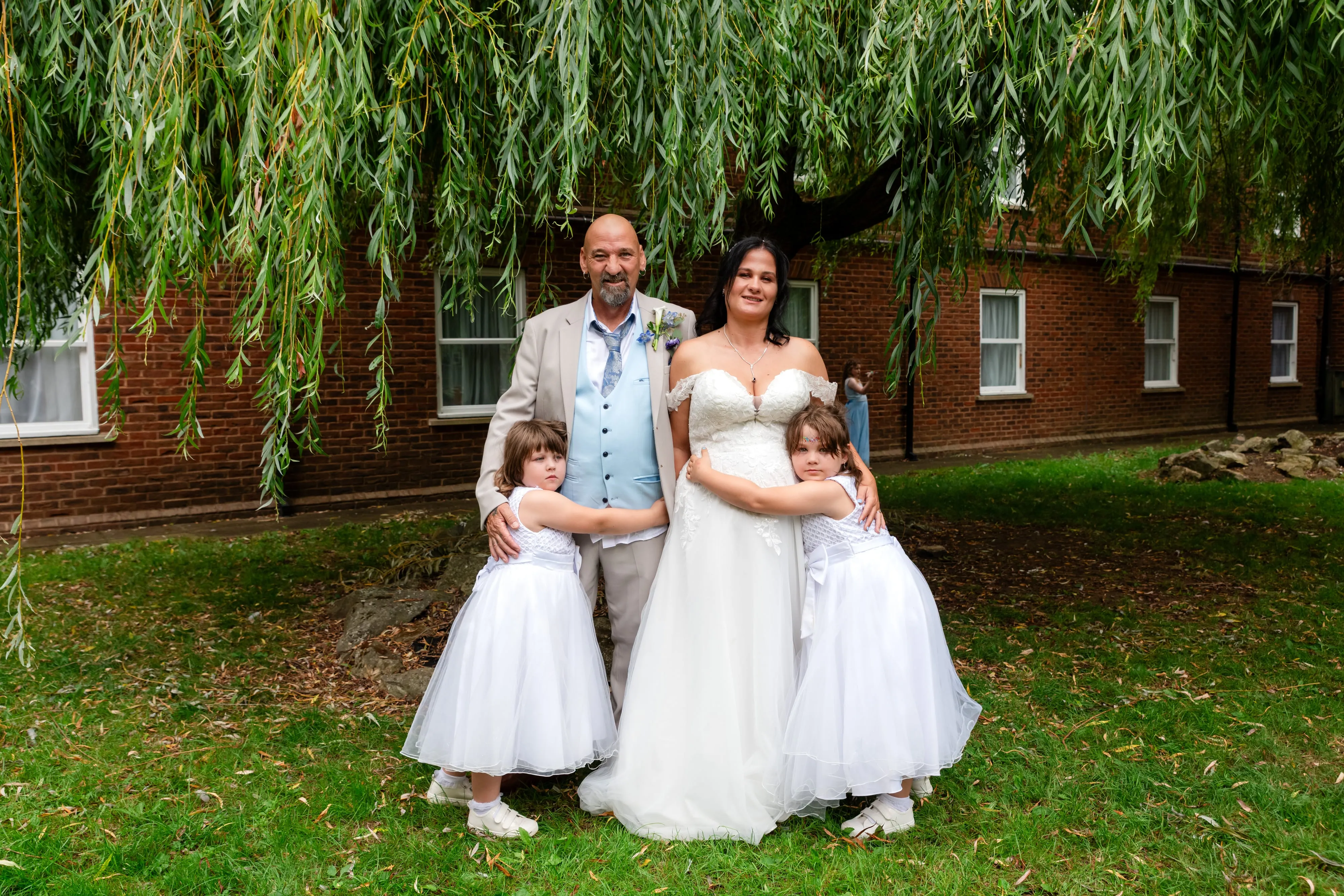 Bride, groom and two flower girls pose under a willow tree in the venue gardens