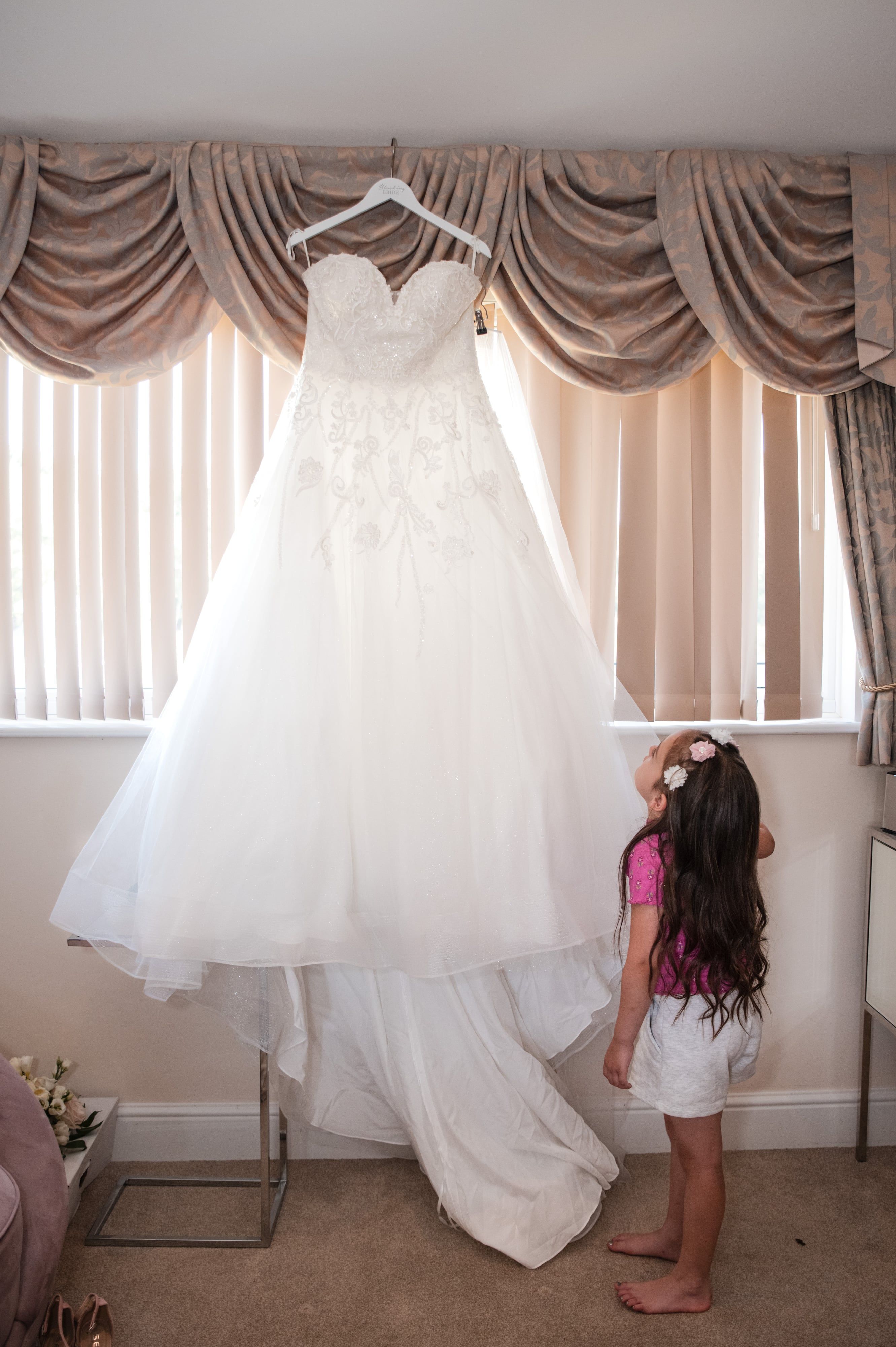 Flower girl reaches up to the wedding dress hanging in the window during bridal preparations