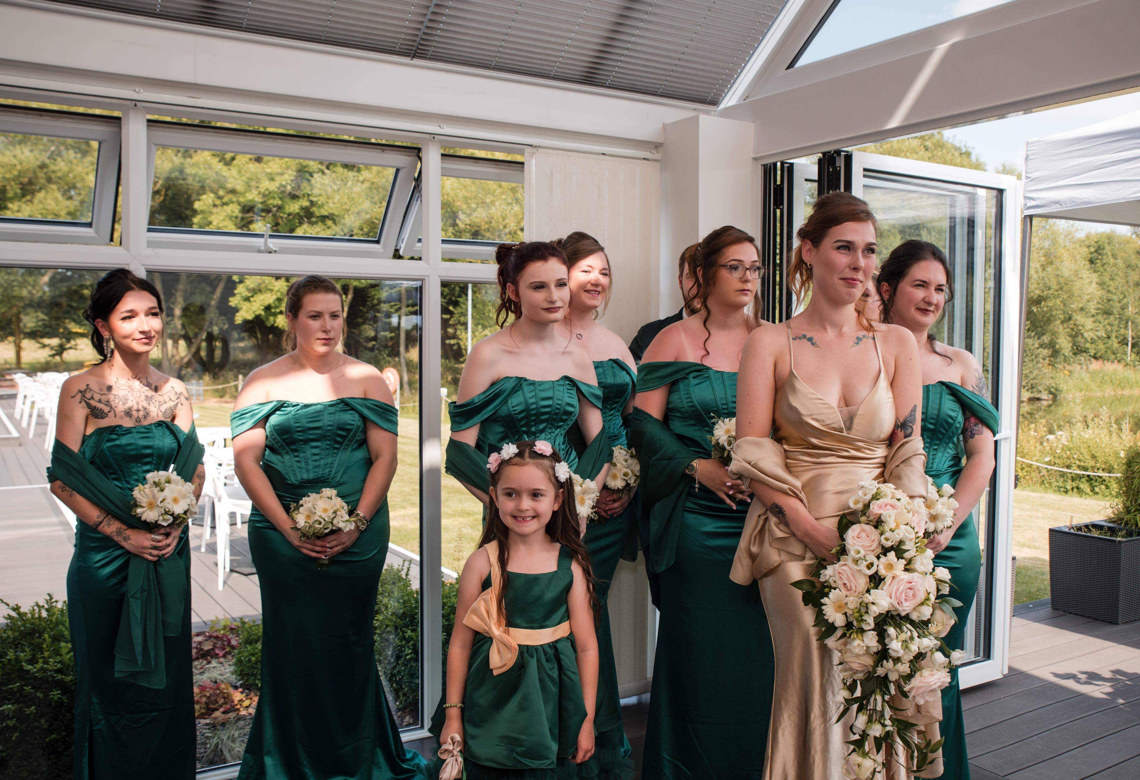 Bridesmaids in emerald green satin wait with the flower girl in the glass ceremony room