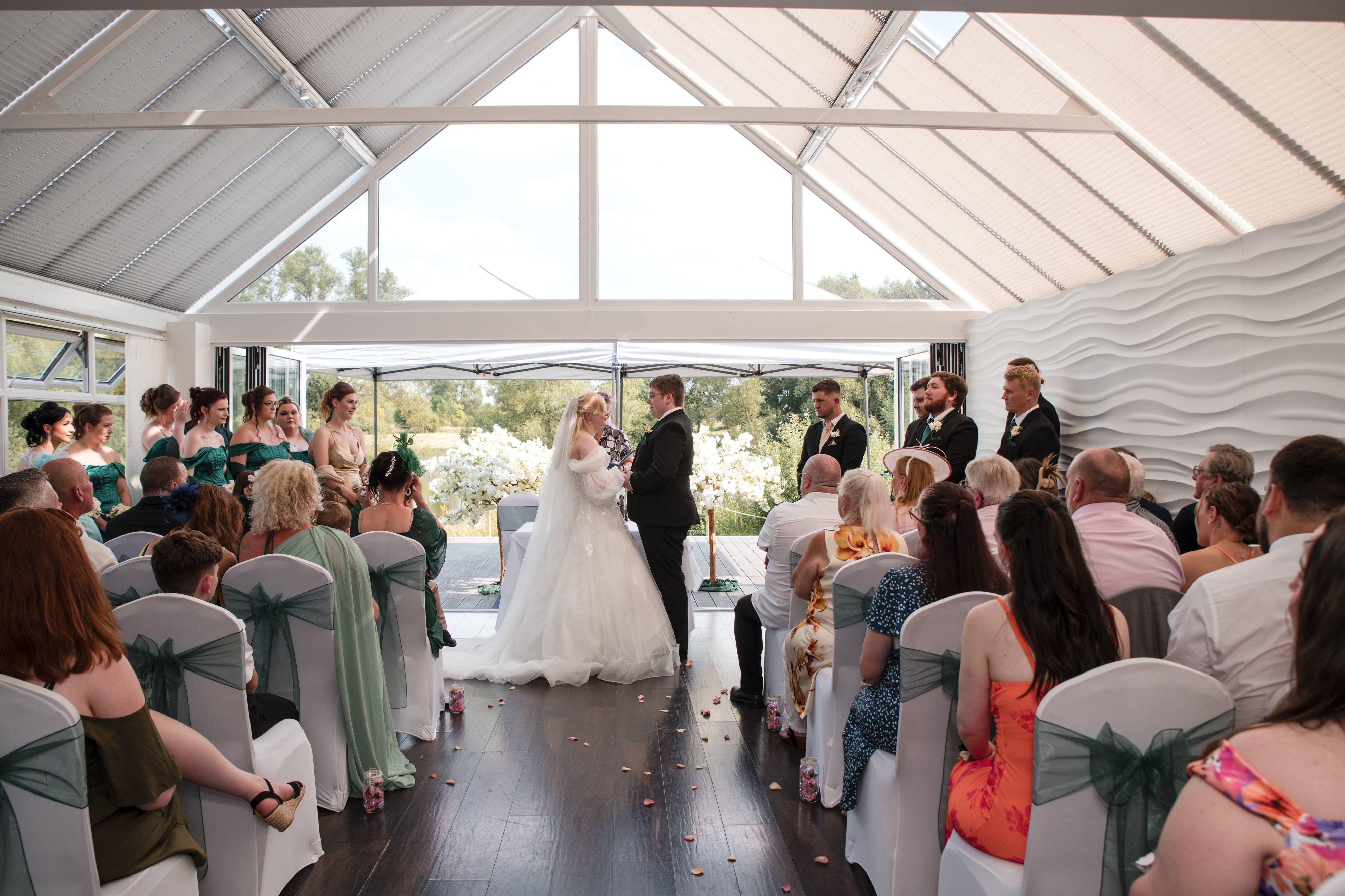 Wide view of the glass-roofed lakeside ceremony with bride, groom and guests