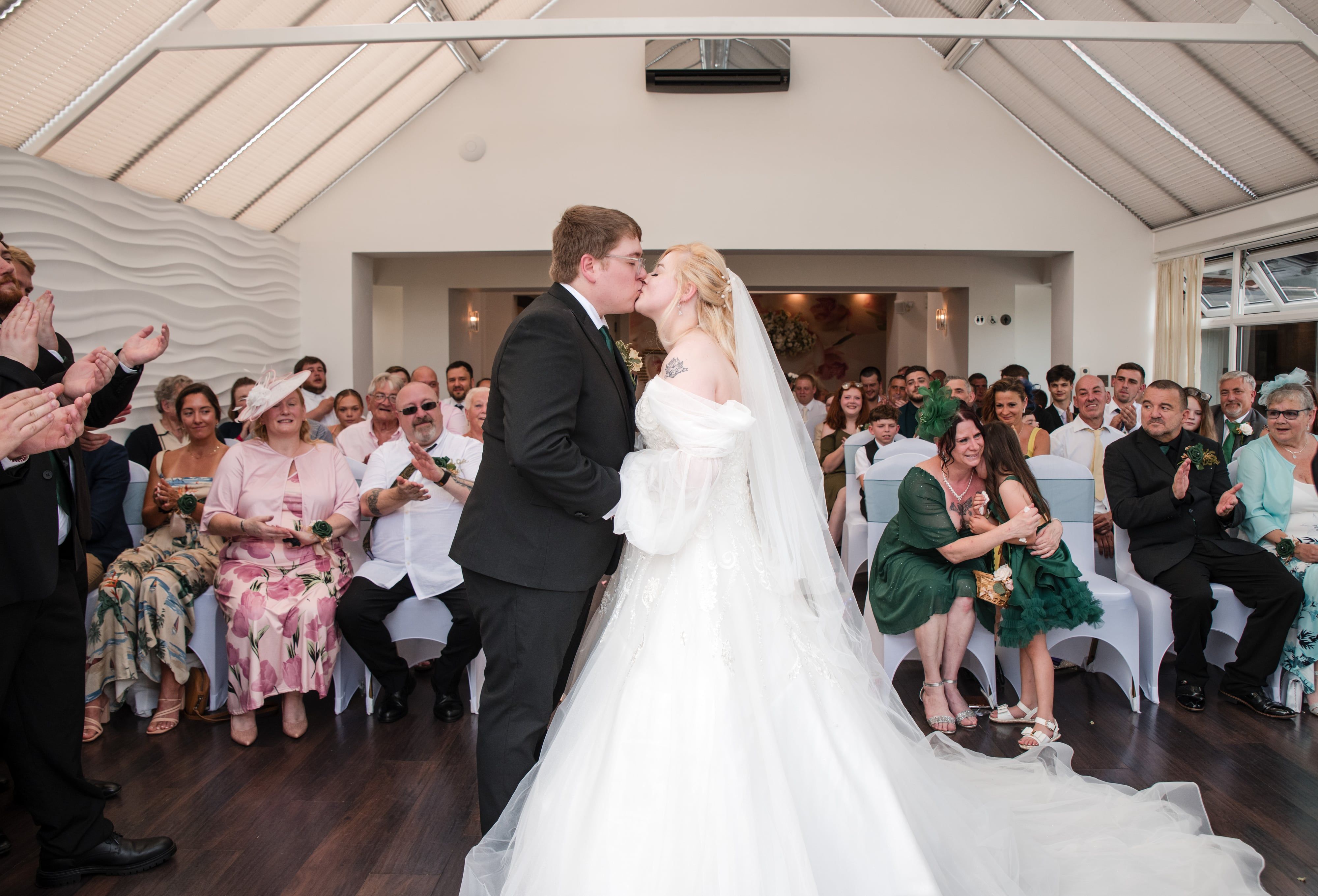 First kiss beneath the pitched glass roof as the wedding party applauds