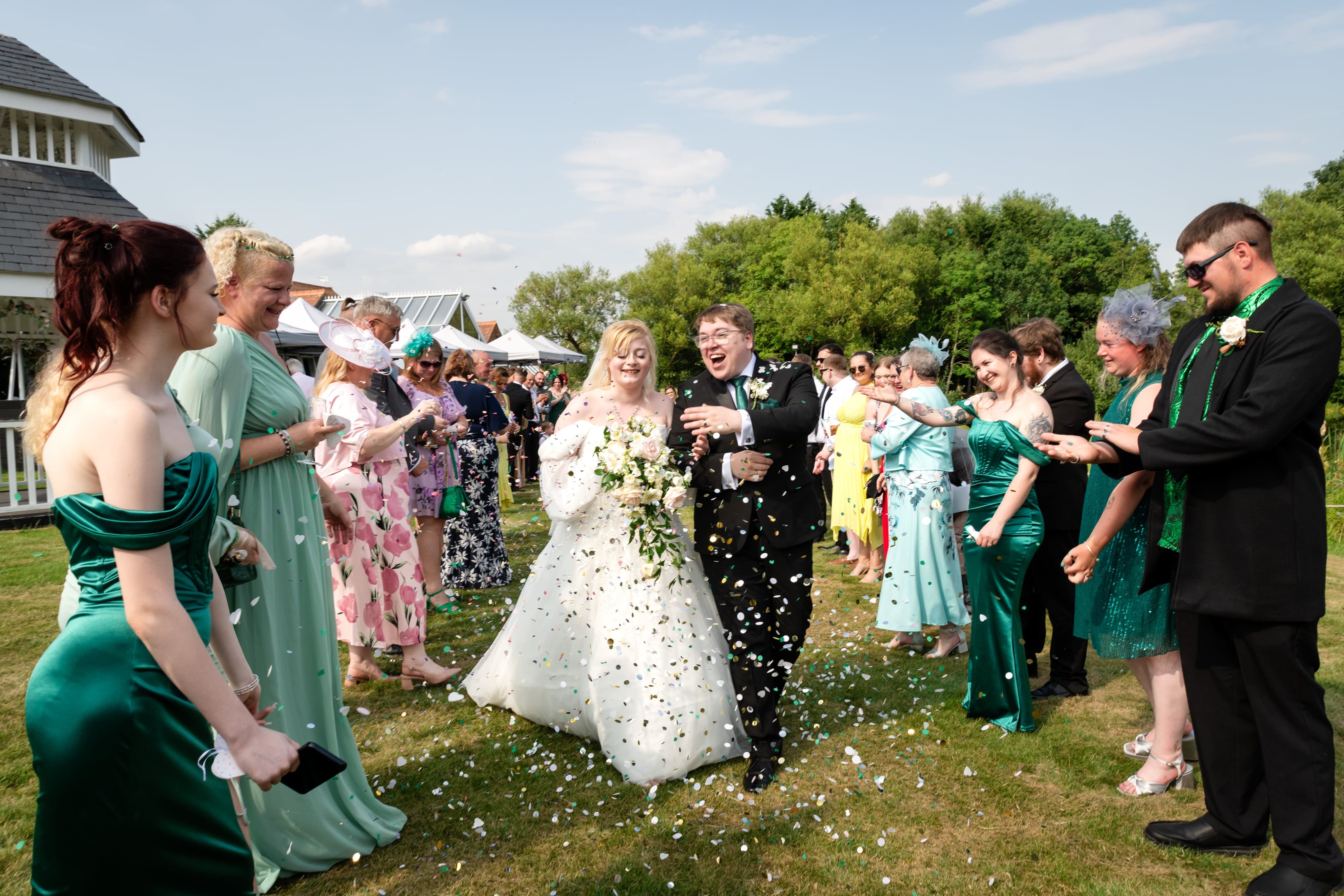 Bride and groom walk through a confetti shower across the Kingfisher lawn, guests lining both sides