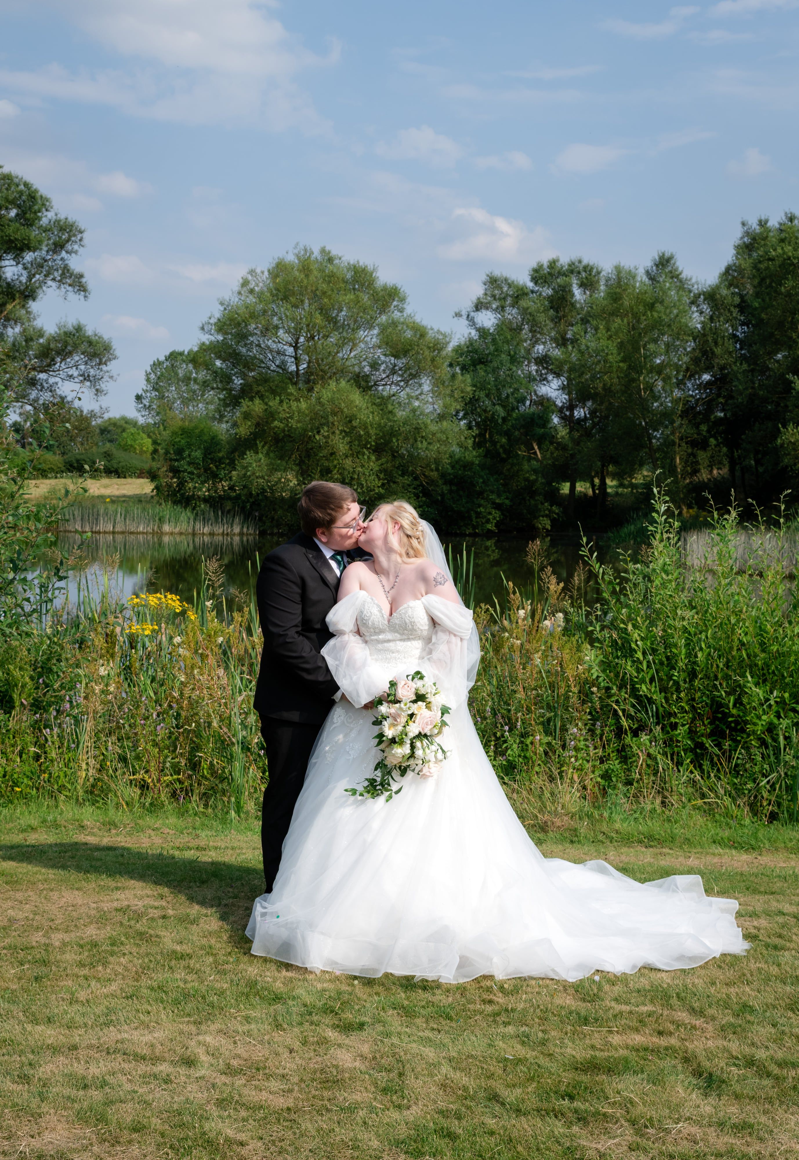 Bride and groom kiss on the lakeside lawn, reeds and water behind them