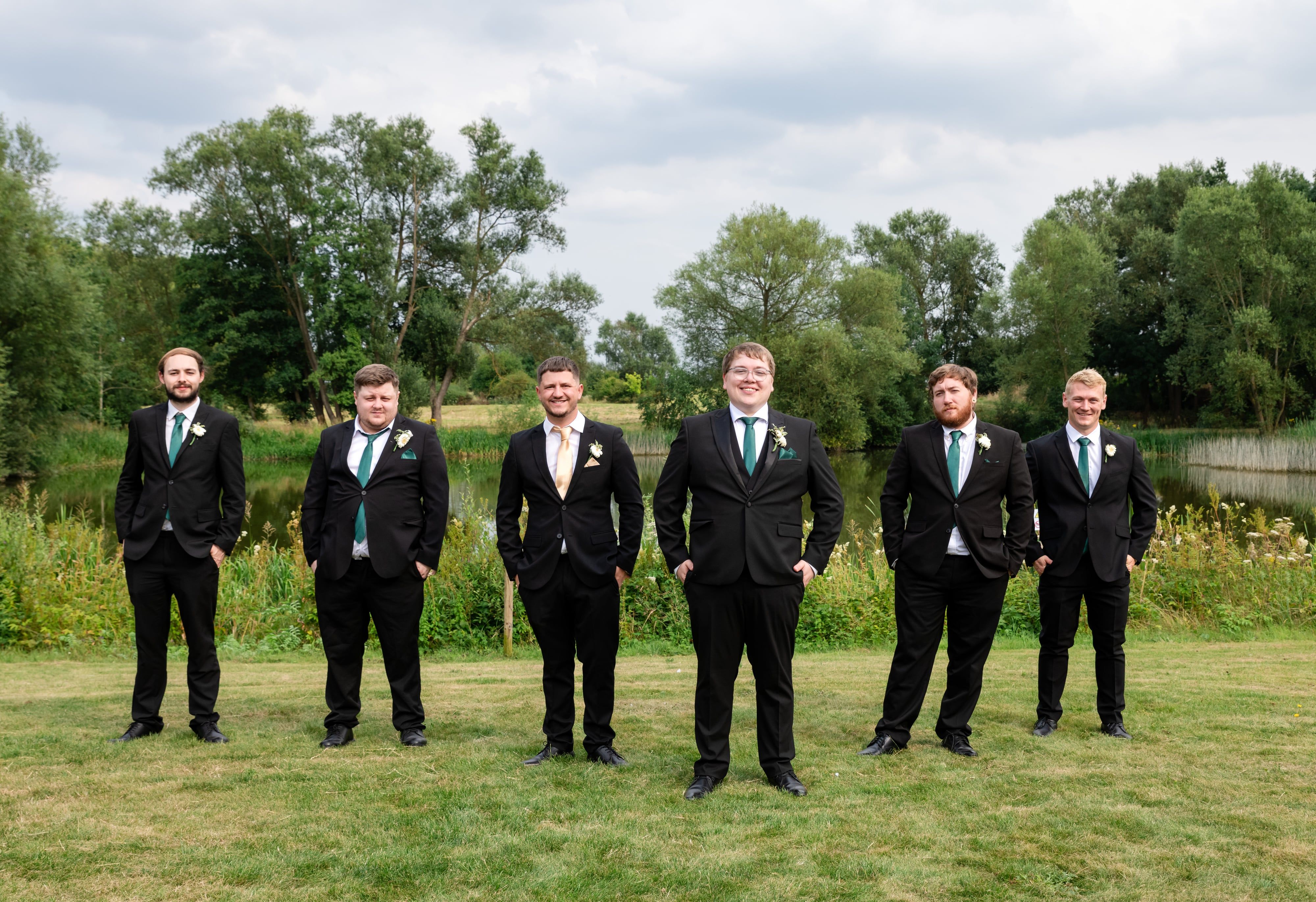 Six groomsmen in dark suits and green ties lined up on the lakeside lawn