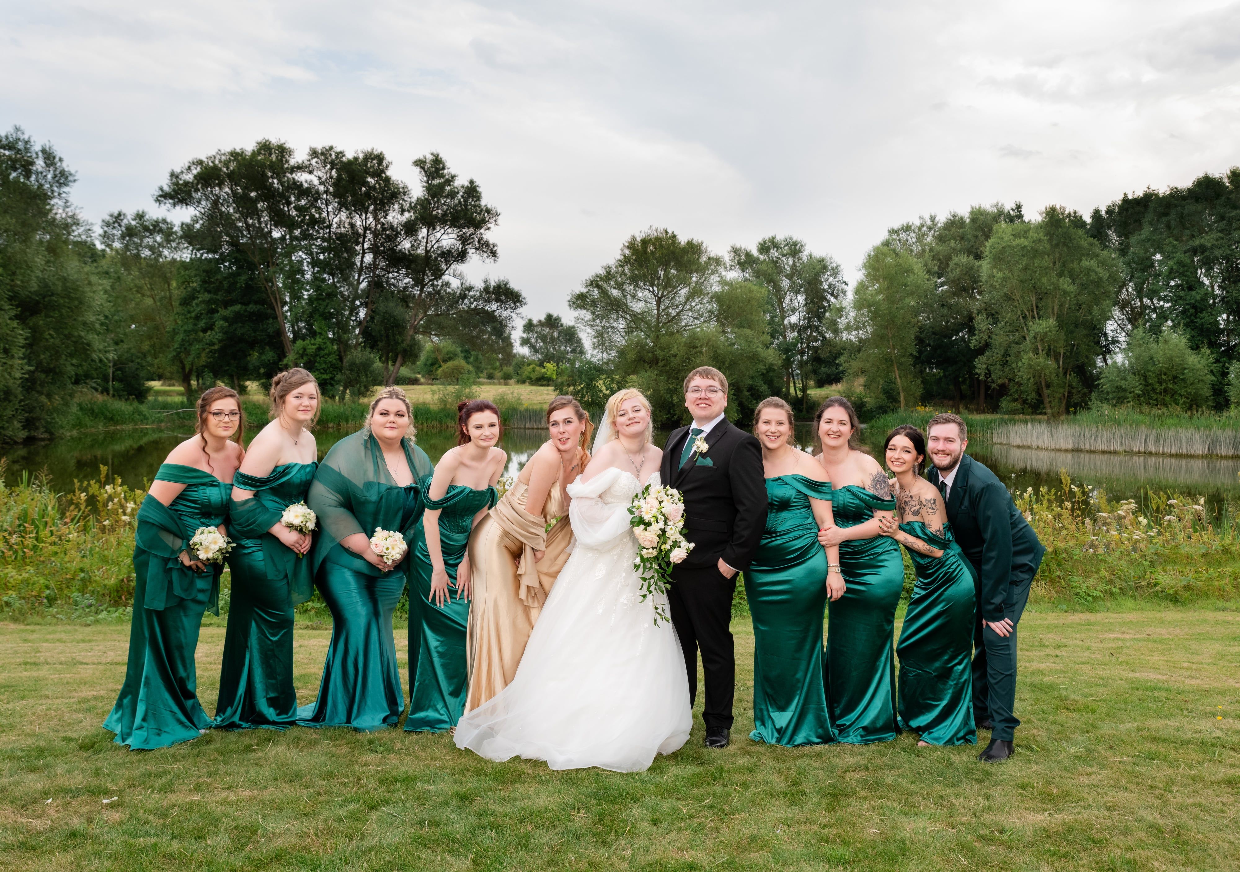Full bridal party in emerald green and gold gathered around the couple by the lake
