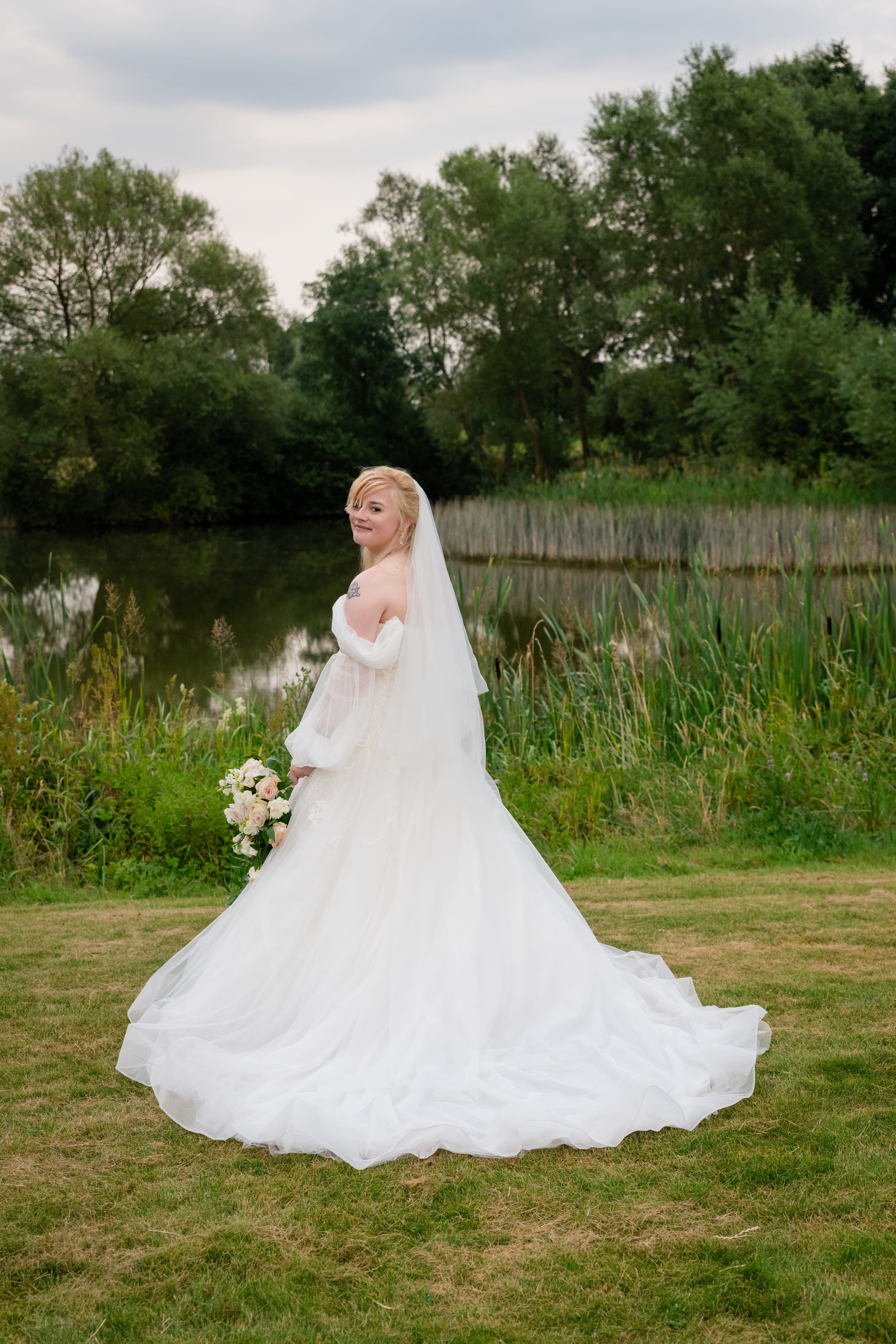 Bride looks over her shoulder in a lakeside portrait, gown sweeping the grass