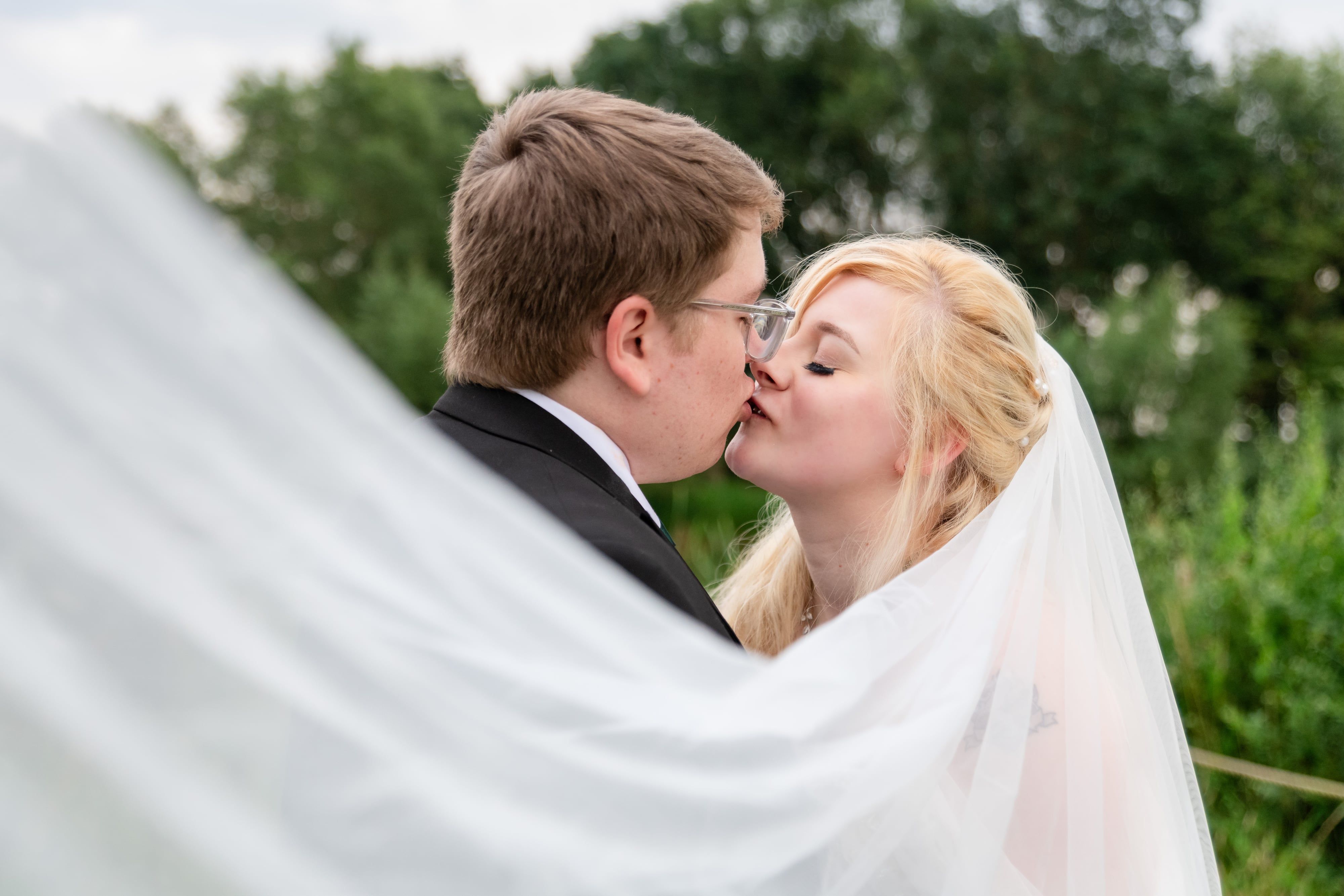 Close-up kiss framed by the sweeping veil against the green of the grounds