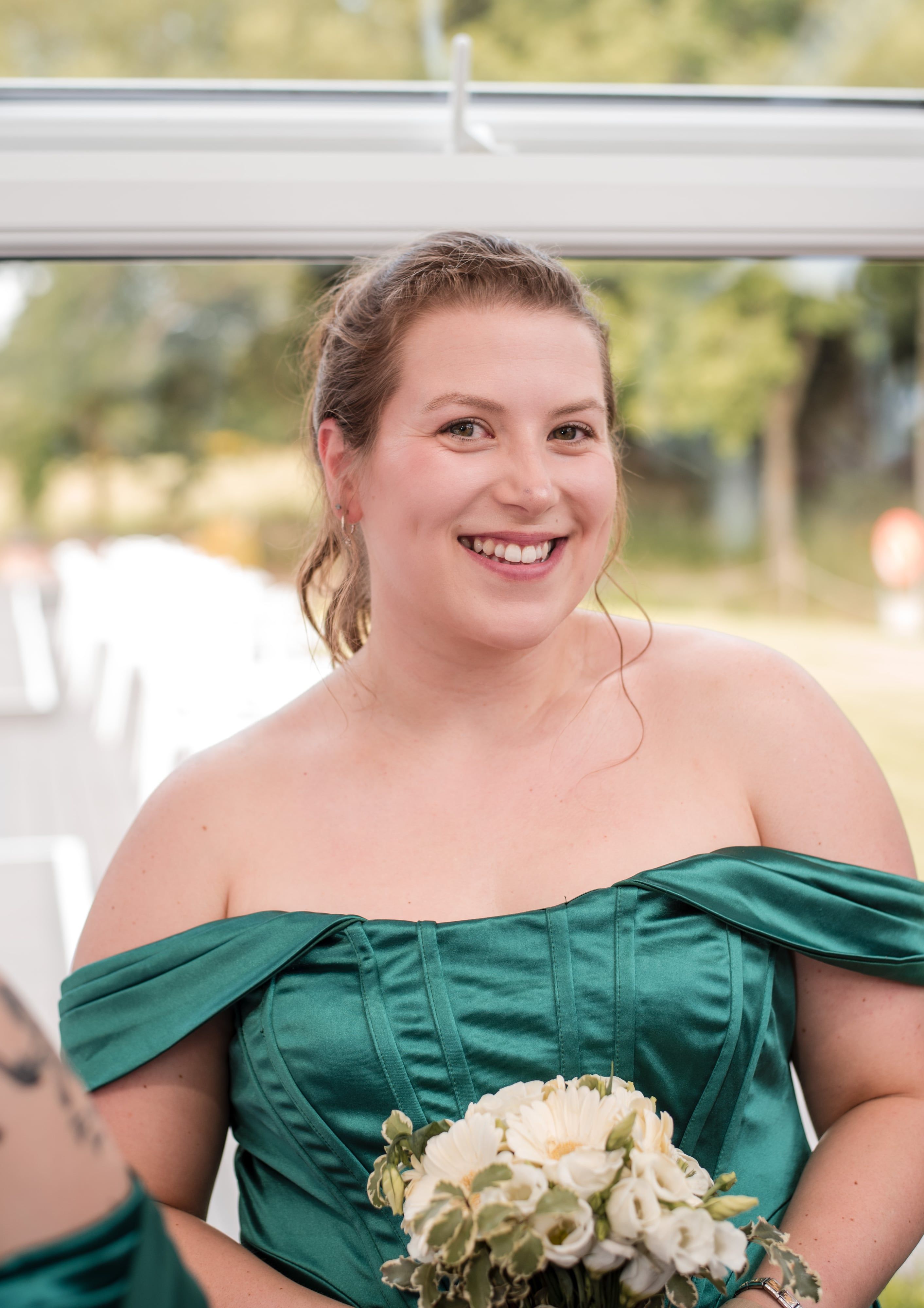 Bridesmaid in emerald green smiles with a white bouquet in the pavilion window
