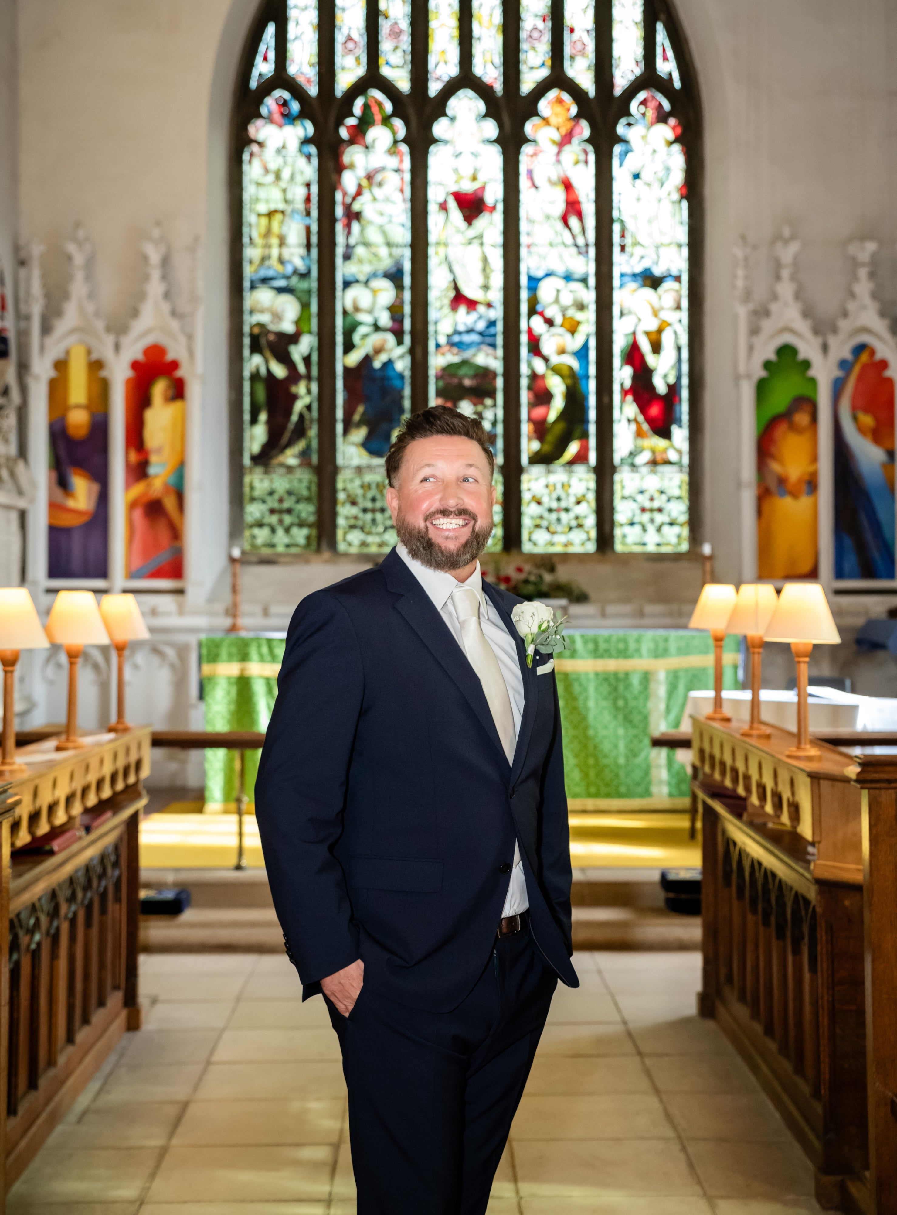 Smiling groom in a navy suit stands before the stained-glass east window at St Andrew's Church, Ampthill