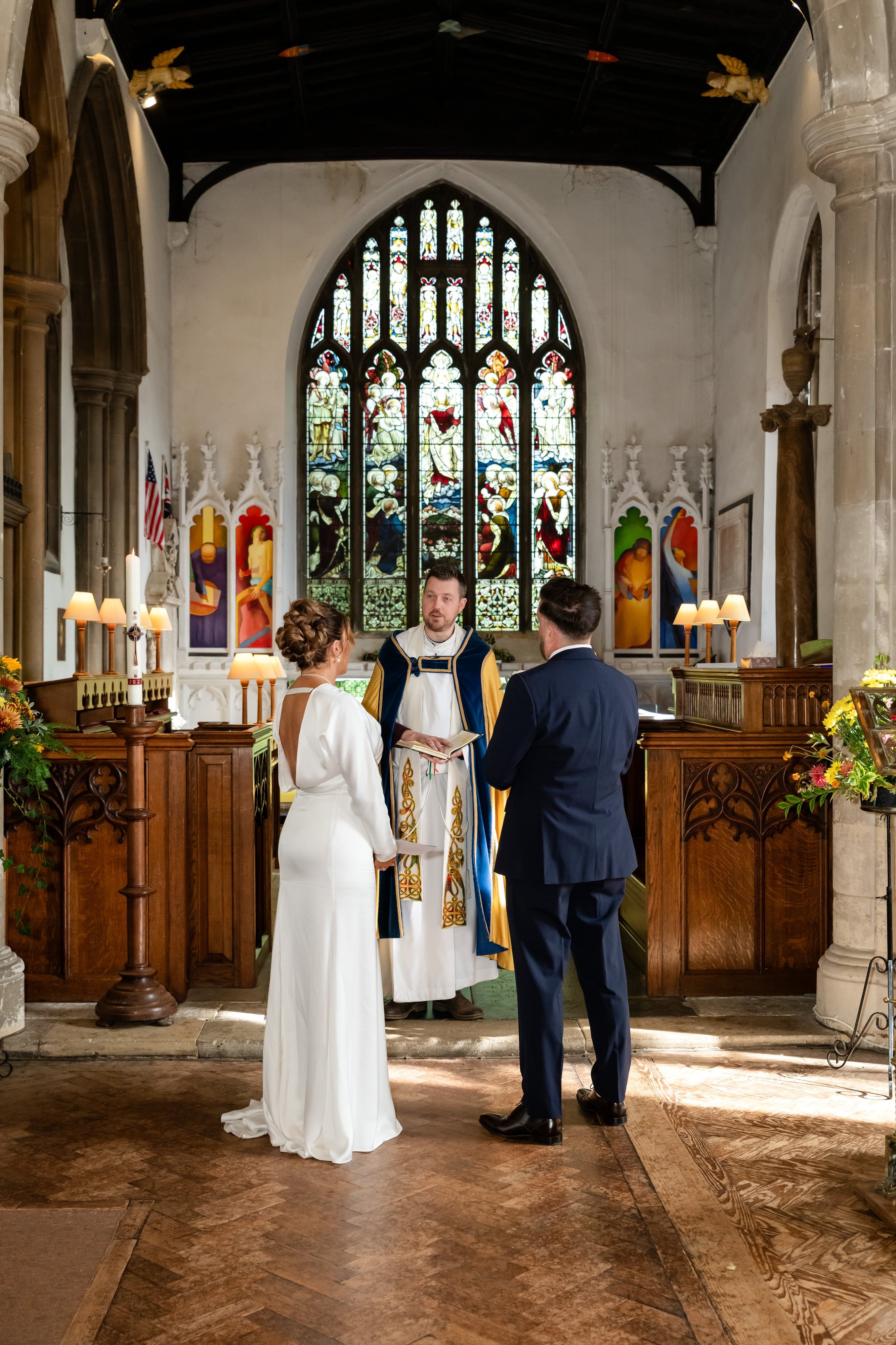 Bride in a low-back satin gown and groom face the vicar at the altar of St Andrew's Church, Ampthill