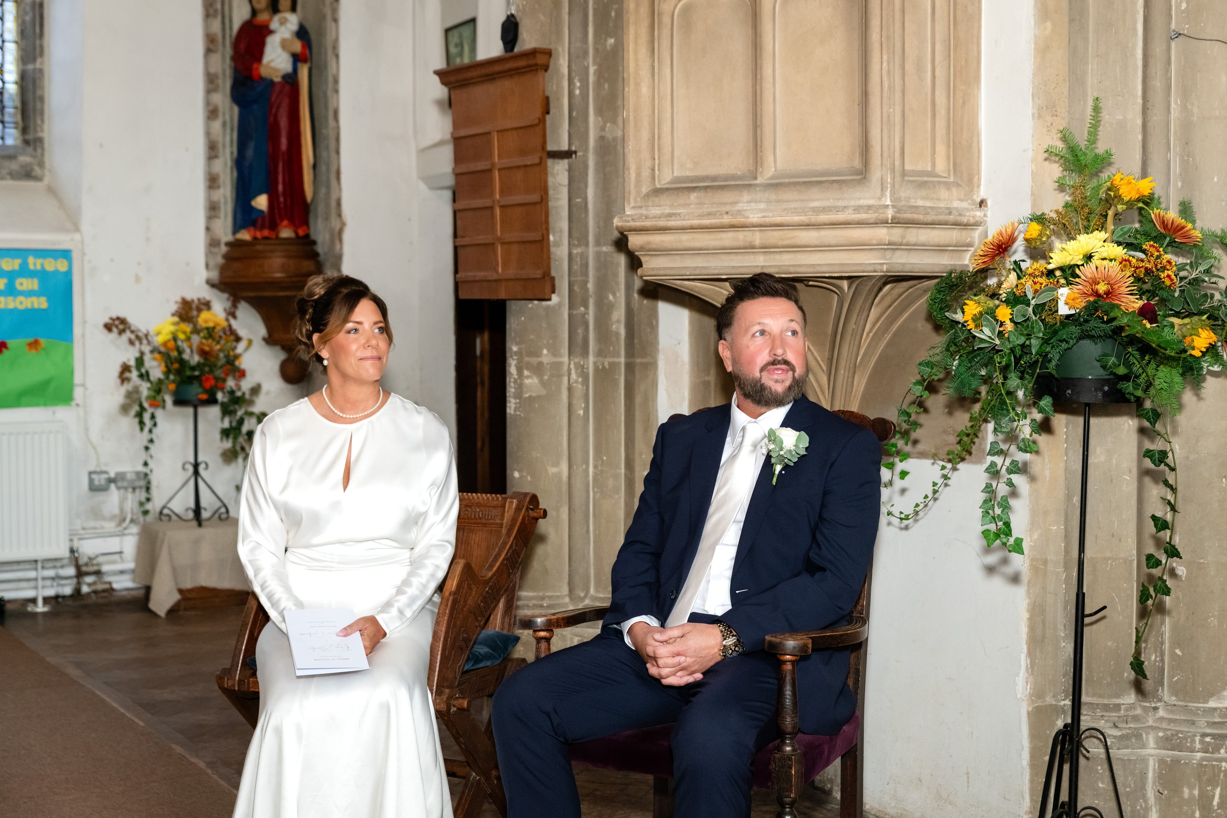 Bride and groom seated side by side during the ceremony, autumn flowers beside the carved stone pulpit
