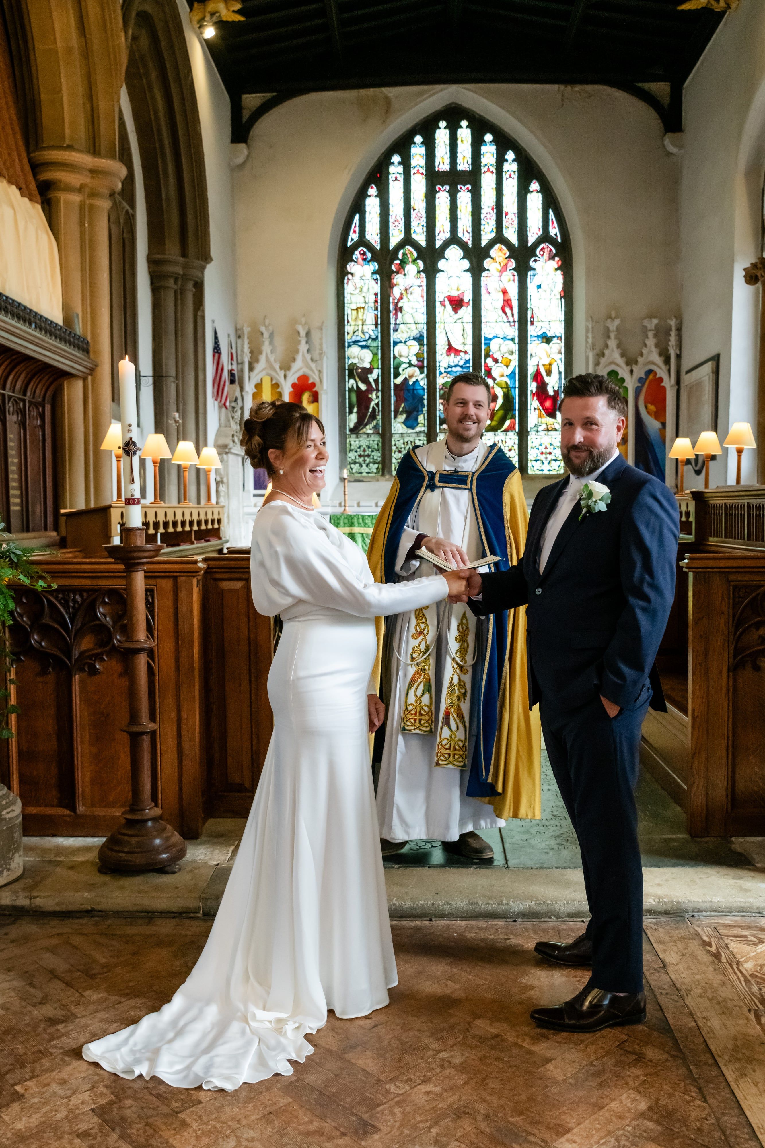 Laughing bride takes the groom's hand during the vows as the vicar smiles beneath the stained glass