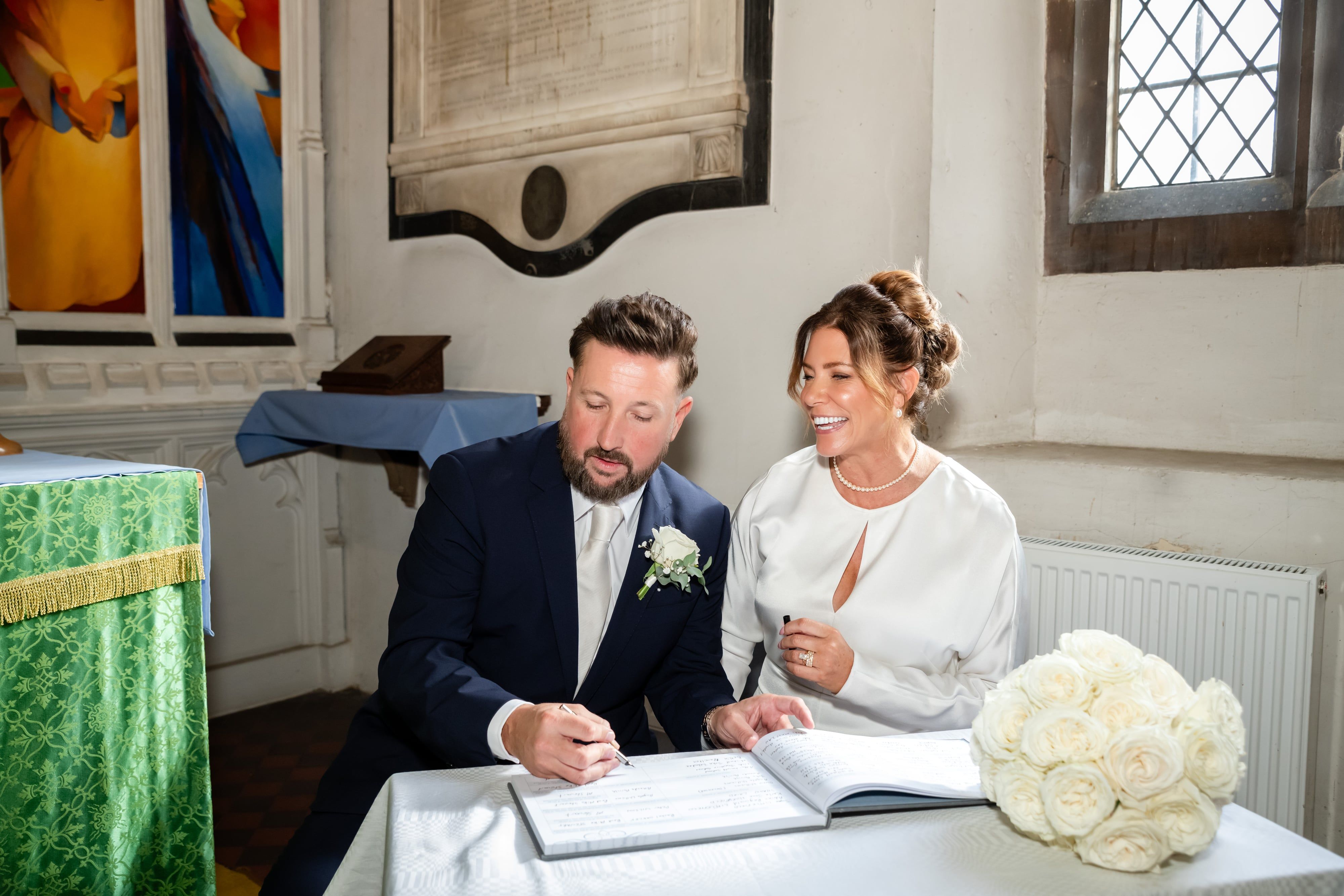 Groom signs the register as the laughing bride looks on, white rose bouquet resting on the table