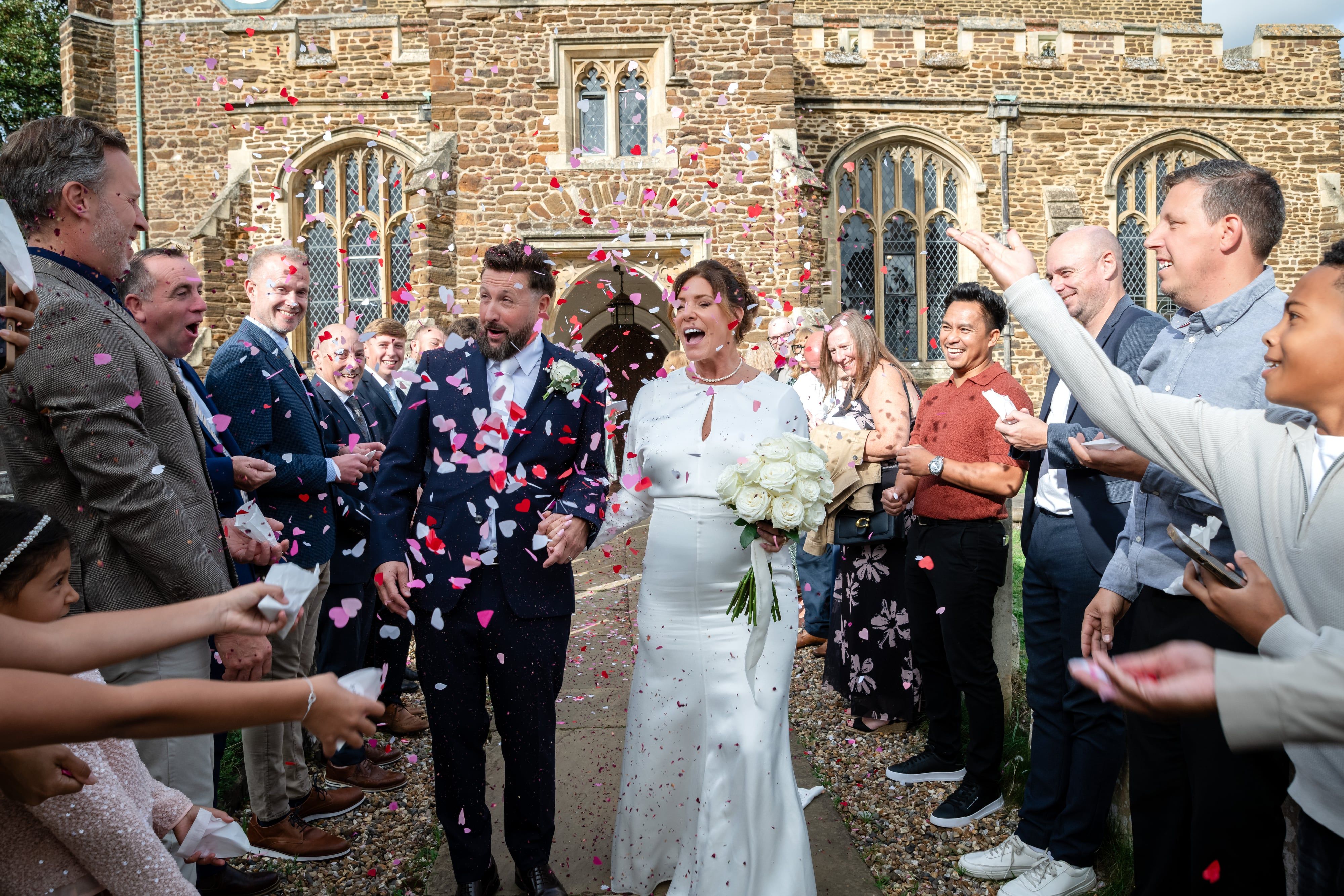 Bride and groom laugh through a shower of pink and red petal confetti outside St Andrew's Church, Ampthill