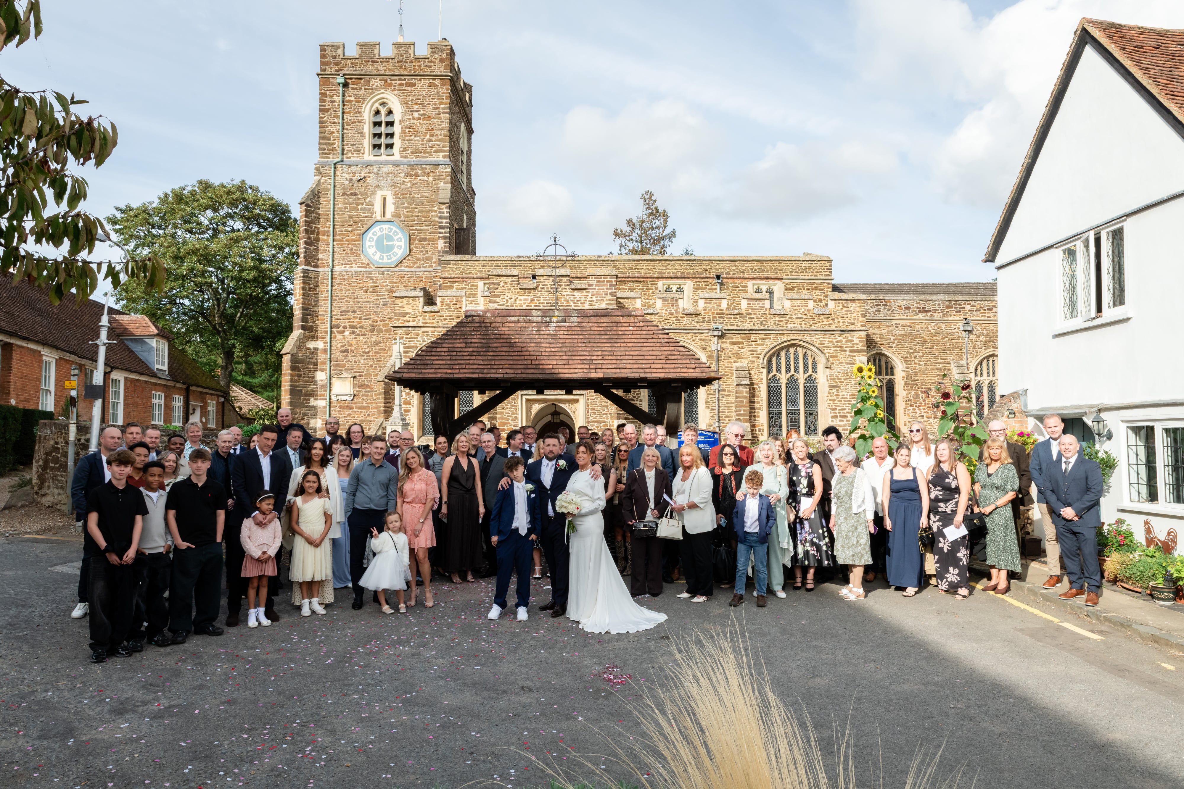 Wedding guests gather with the couple beneath the clock tower and lychgate of St Andrew's, Ampthill