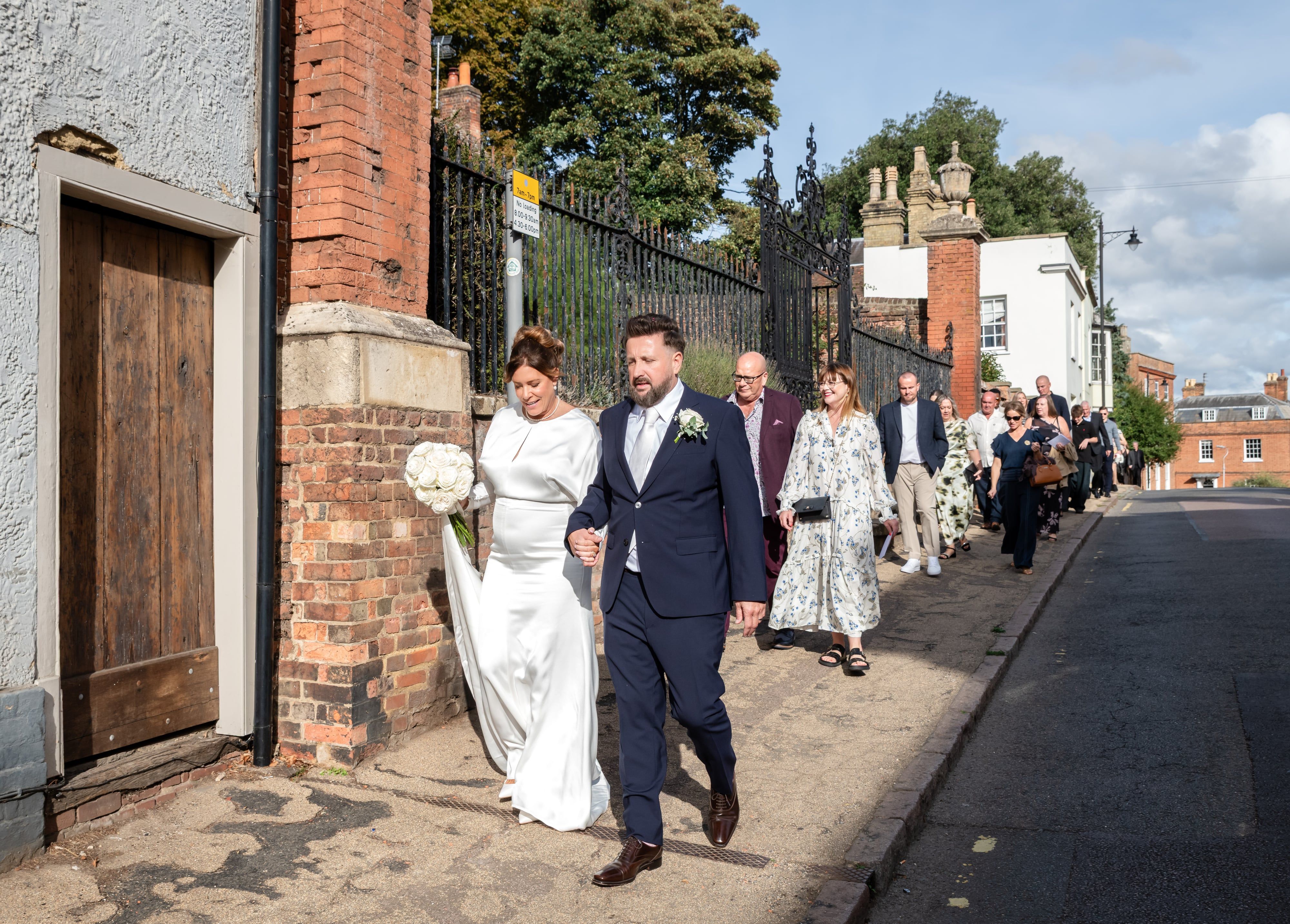 Newlyweds lead their guests along a brick-walled Ampthill street past wrought-iron gates