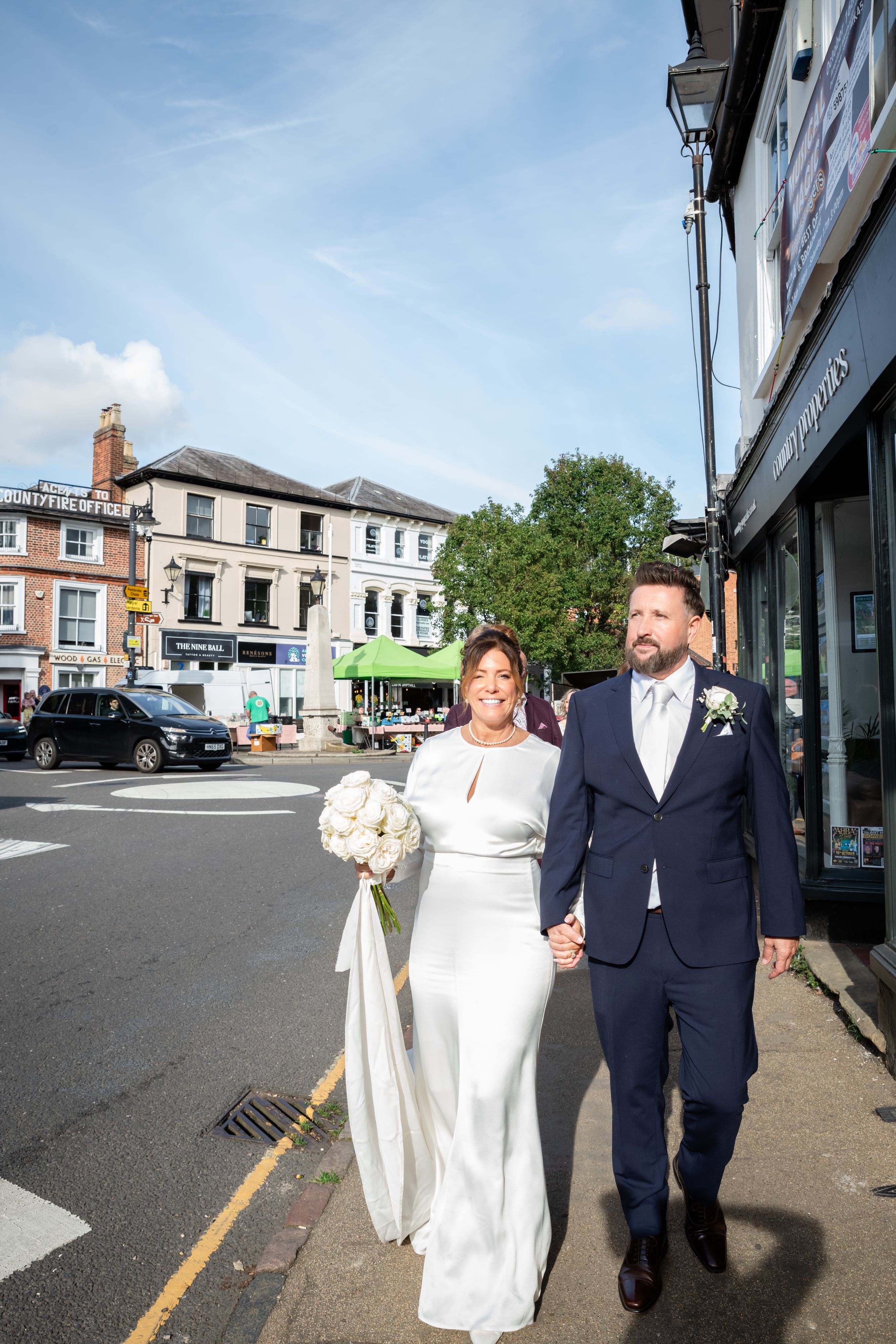 Smiling bride and groom walk hand in hand through Ampthill town centre in the sunshine
