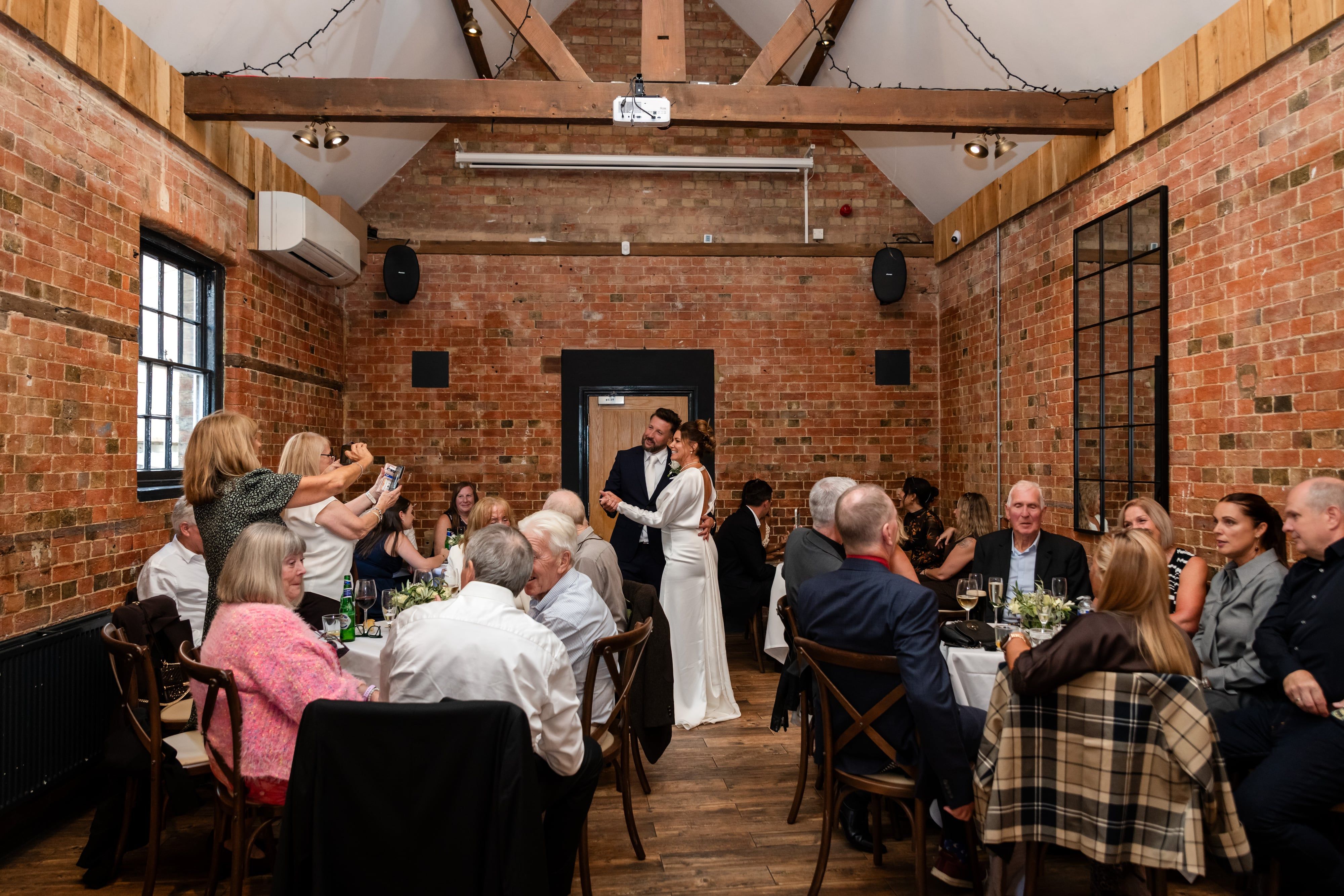Bride and groom share a dance between tables in an exposed-brick reception room as guests film on their phones