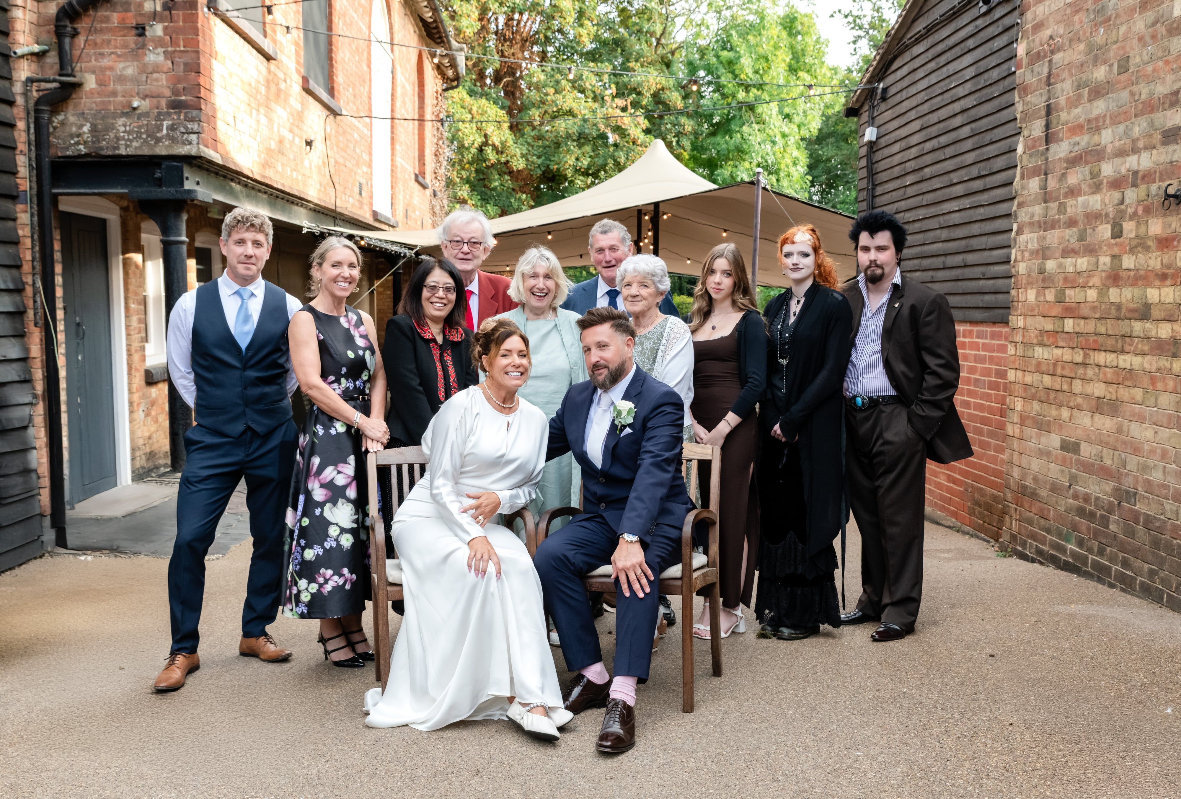 Newlyweds seated with family in a brick courtyard beneath a sail canopy and festoon lights