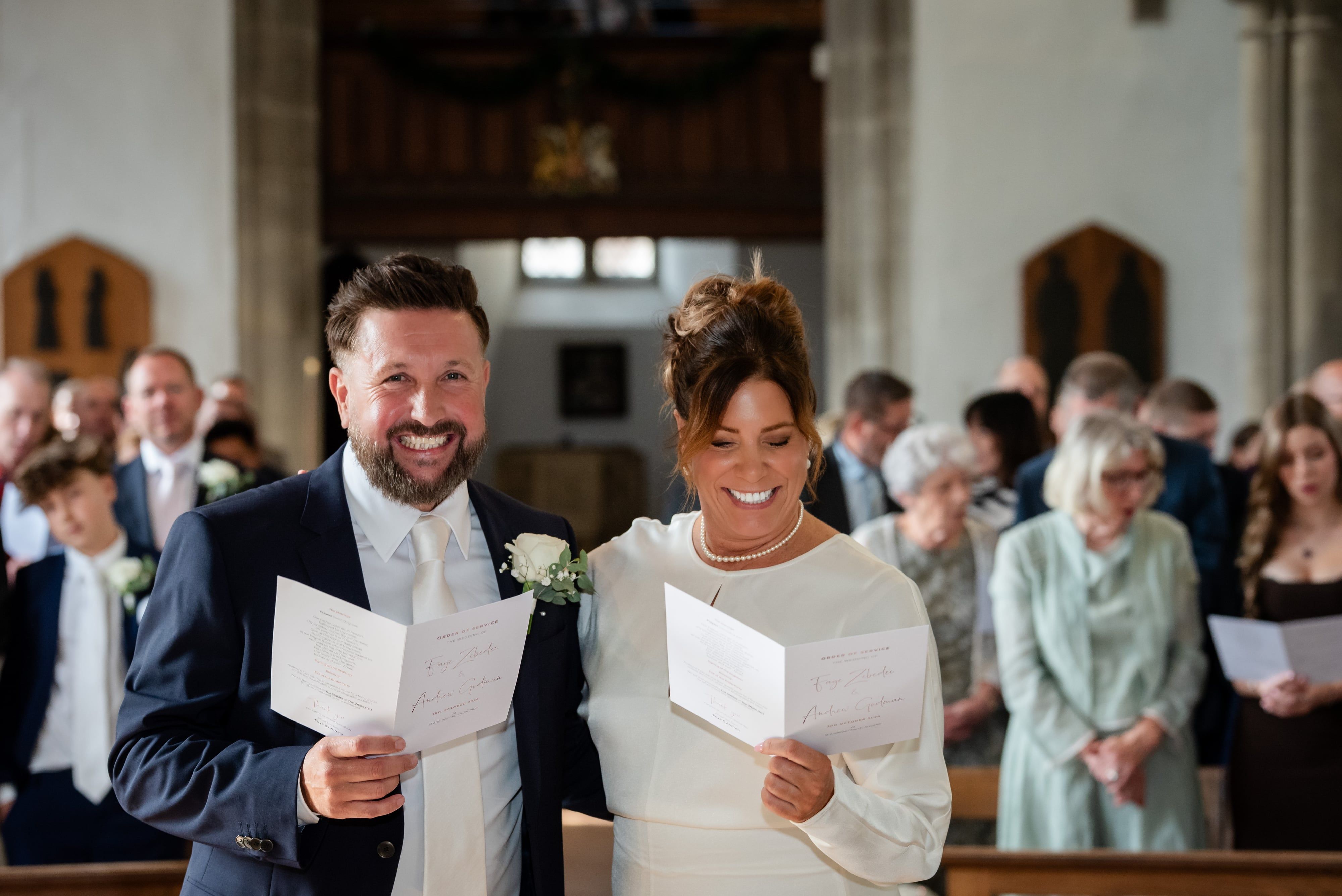 Bride and groom laugh as they sing from their orders of service in St Andrew's Church, Ampthill