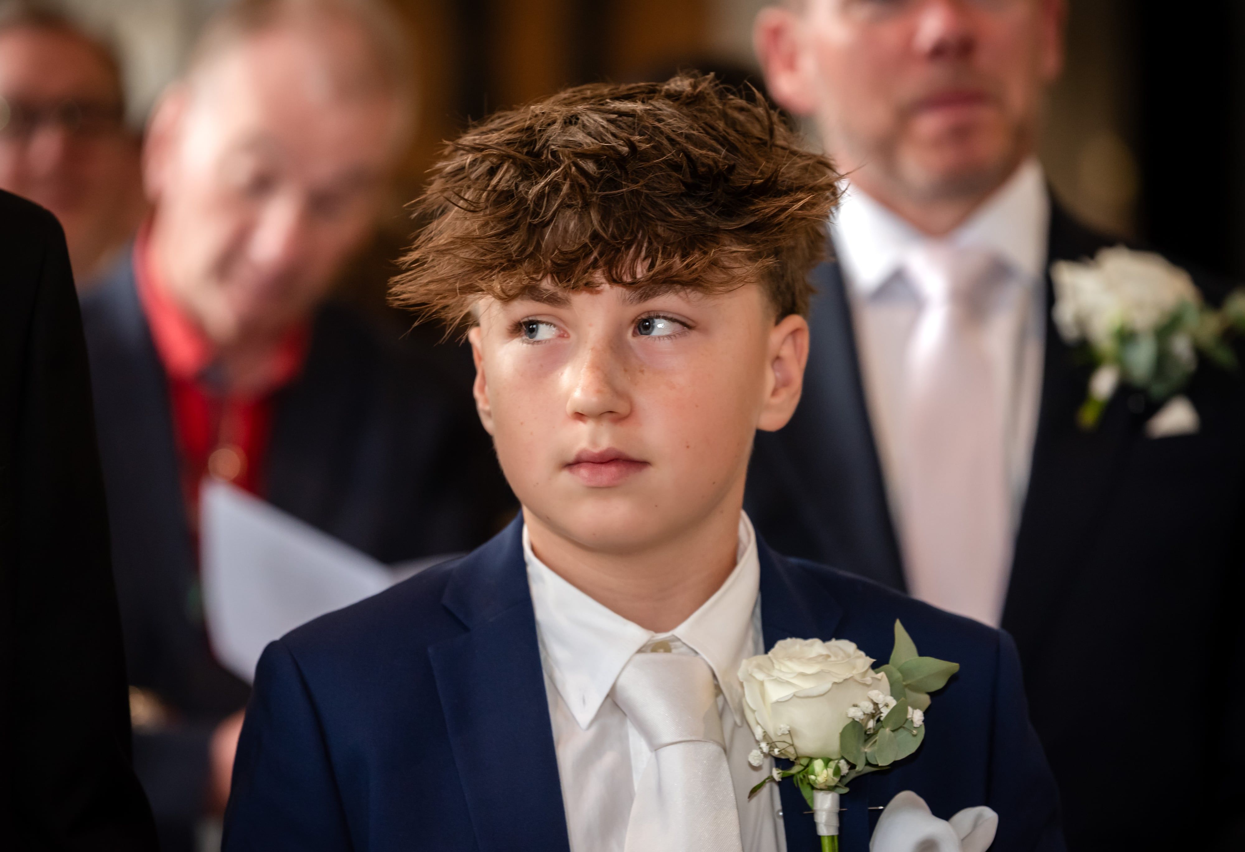 Young guest in a navy suit and white rose buttonhole glances across the church during the ceremony
