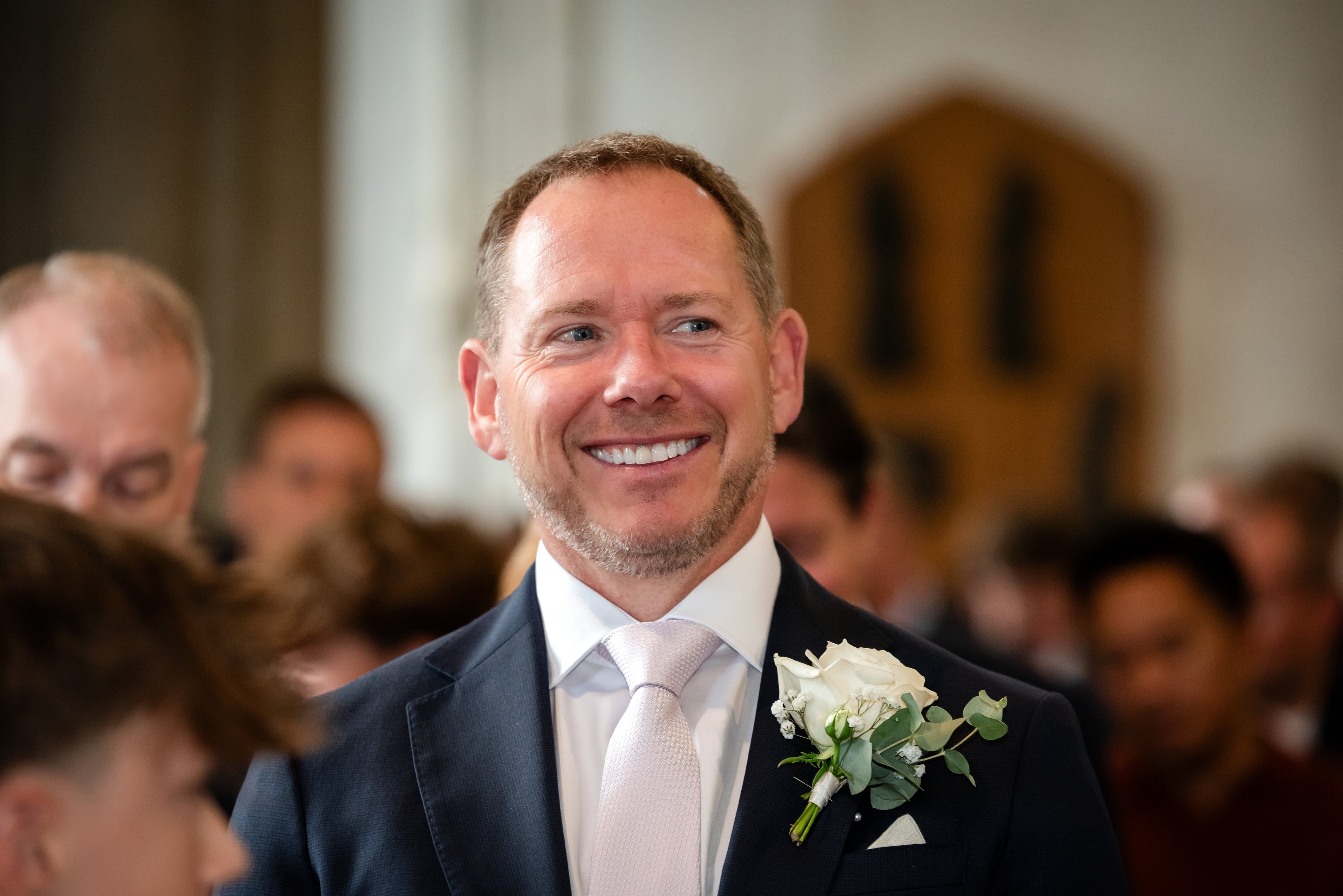 Smiling guest in a navy suit with white rose buttonhole and pale pink tie watches the wedding ceremony
