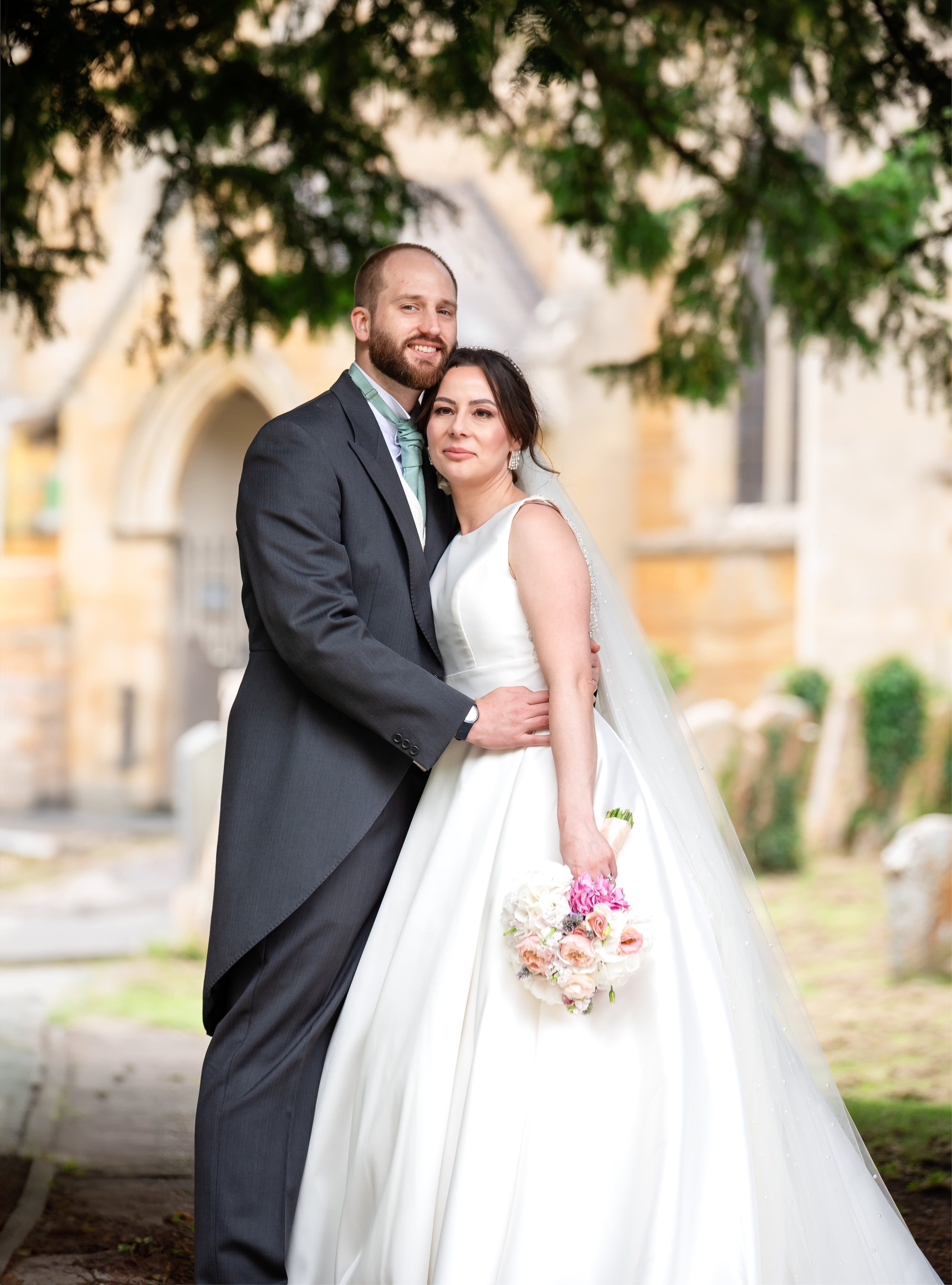 Bride and groom embrace in the churchyard beneath an evergreen, ironstone church behind