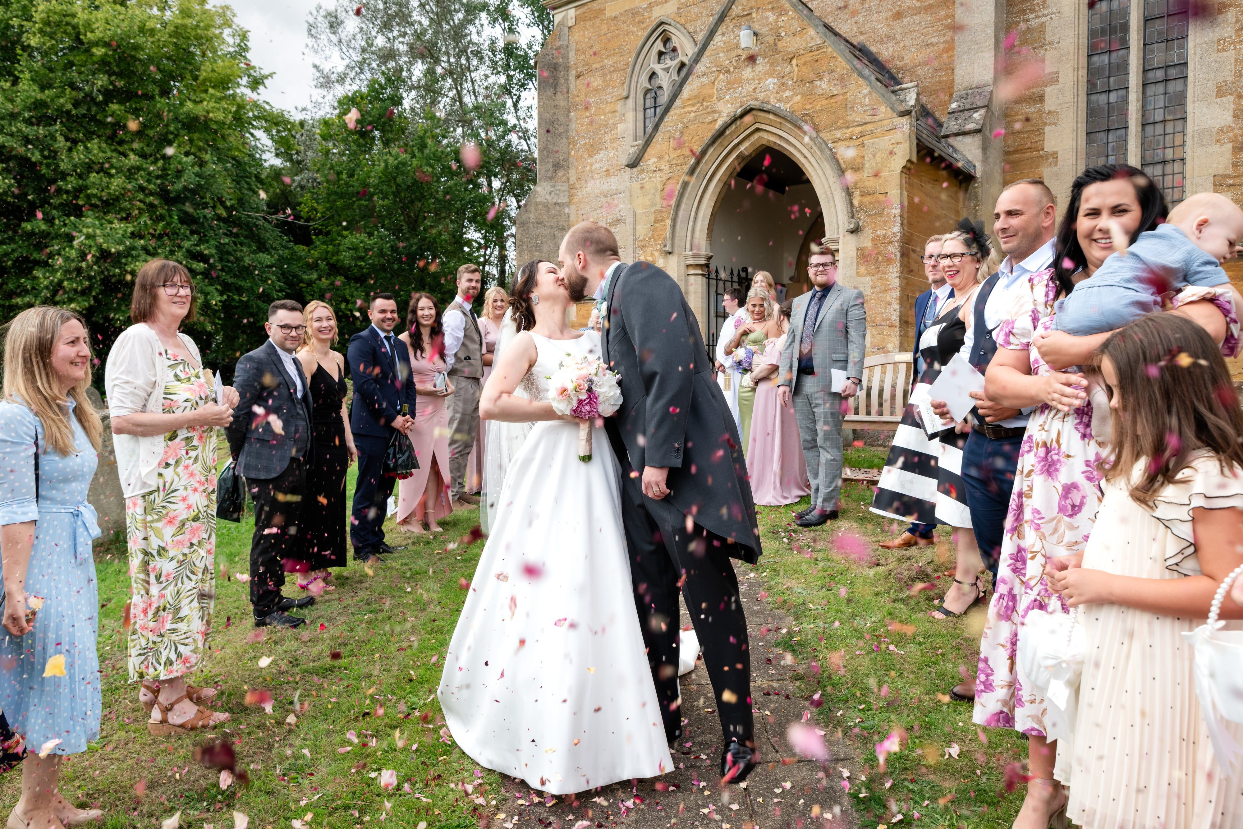 Bride and groom kiss in a confetti shower on the green outside St Mary's Church, Orlingbury
