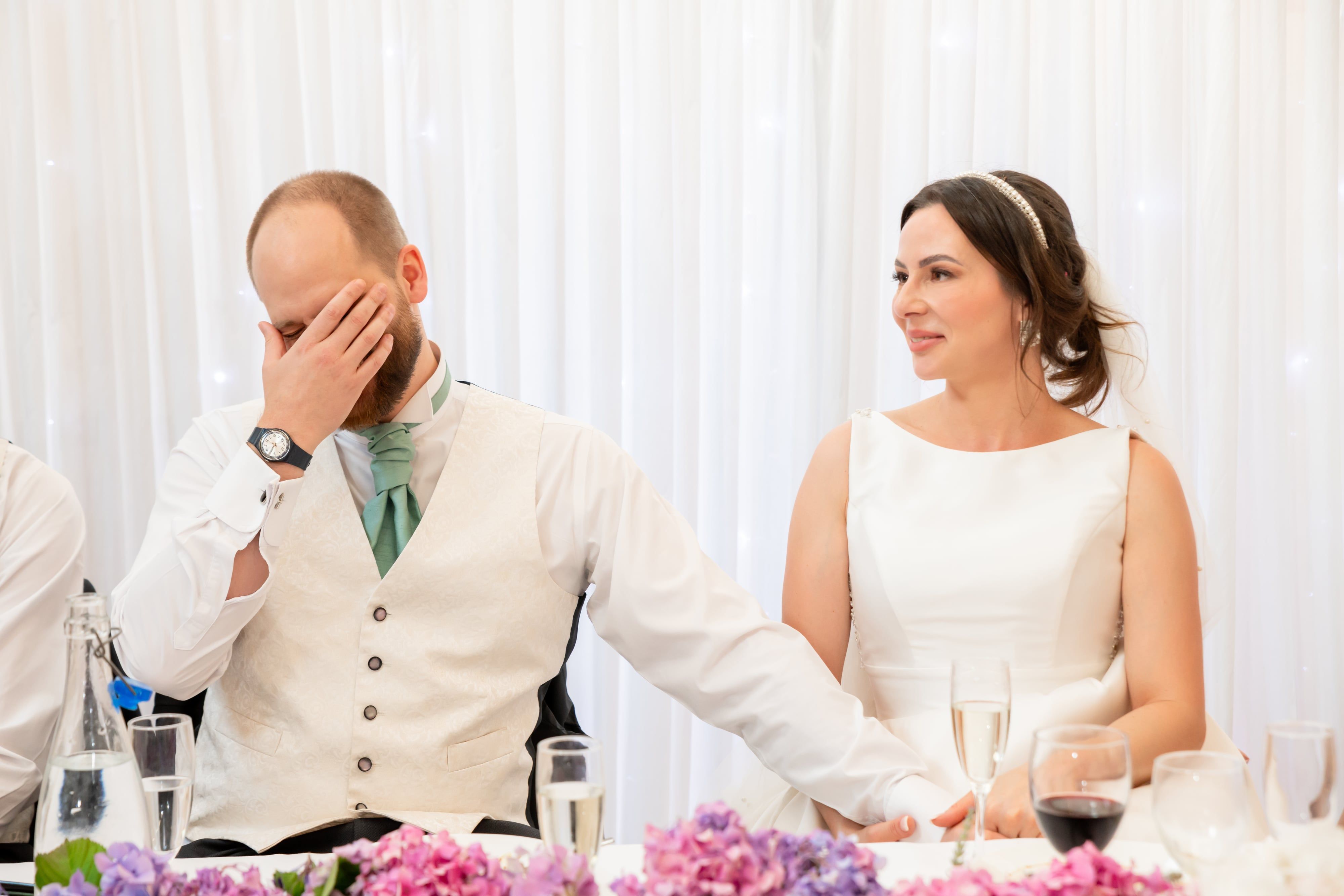 Groom laughs into his hand during the best man speech as the bride looks on