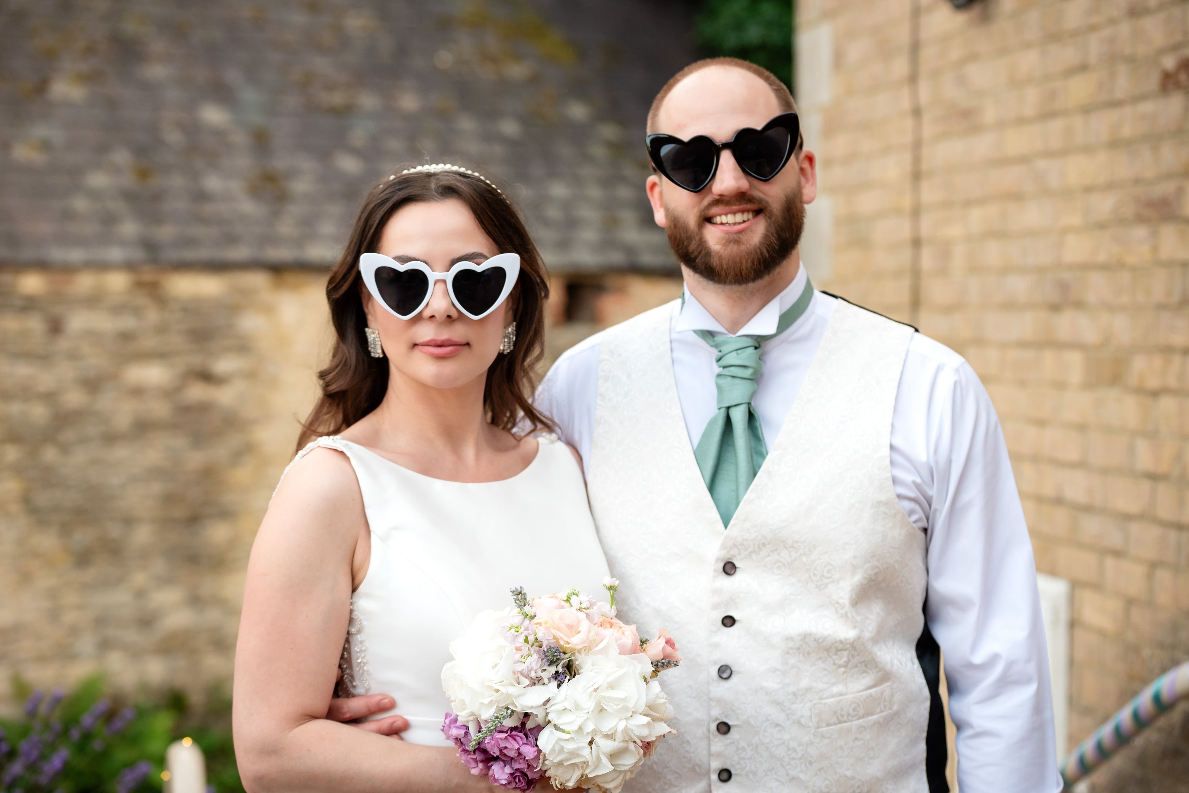 Bride and groom in heart-shaped sunglasses grin outside the reception